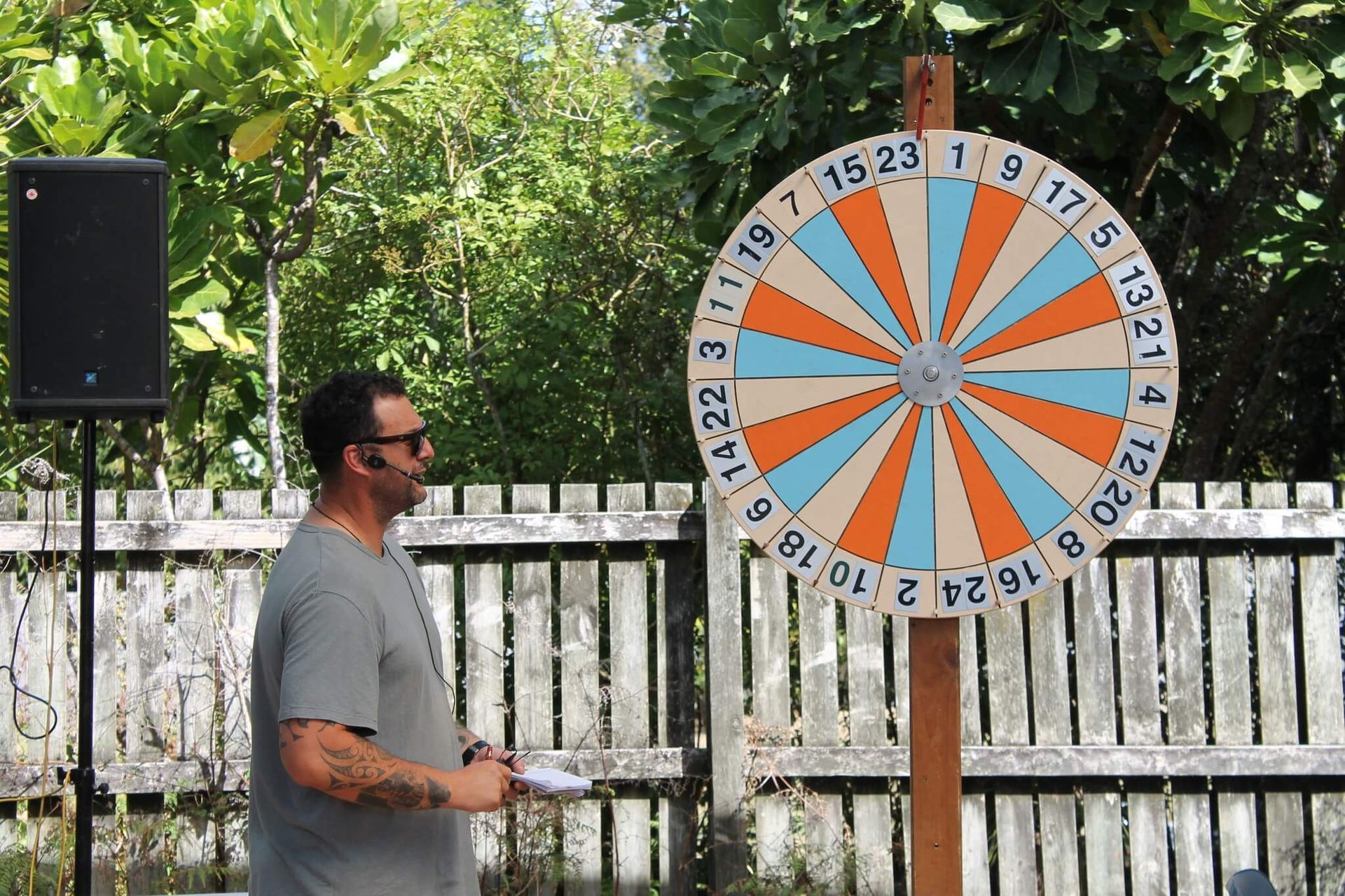 Man spinning a large game wheel with numbers. Outdoors, near a fence, speaker on the left.