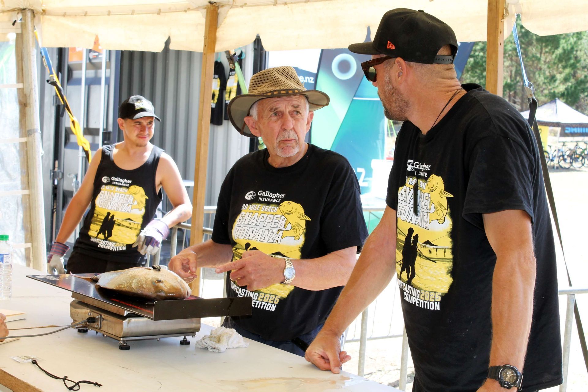 Three people at a counter under a tent. Two men examine food on a scale. Another man stands behind the counter.