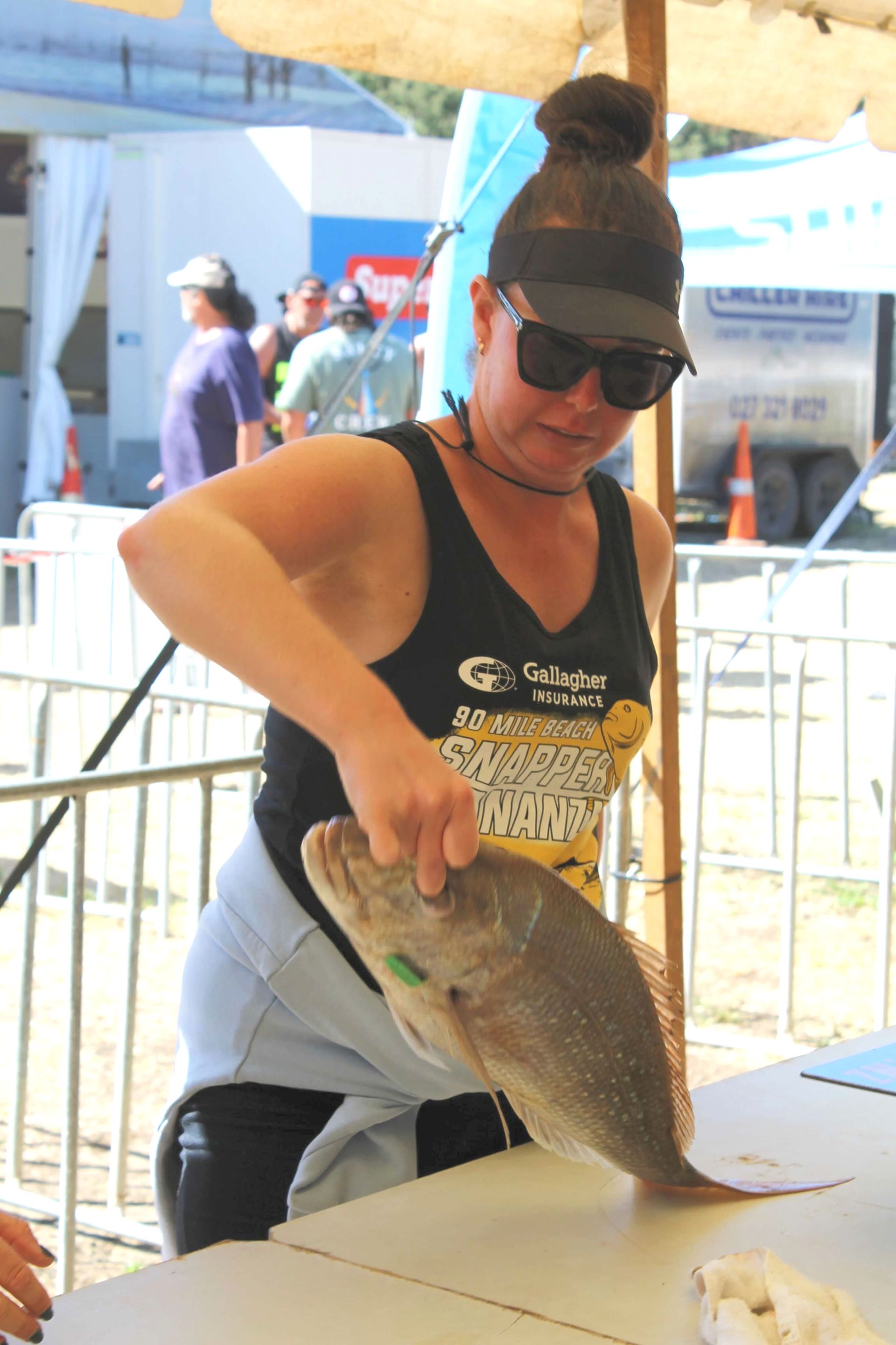 Woman holding a fish at a table, wearing sunglasses, black tank top, outdoor setting.