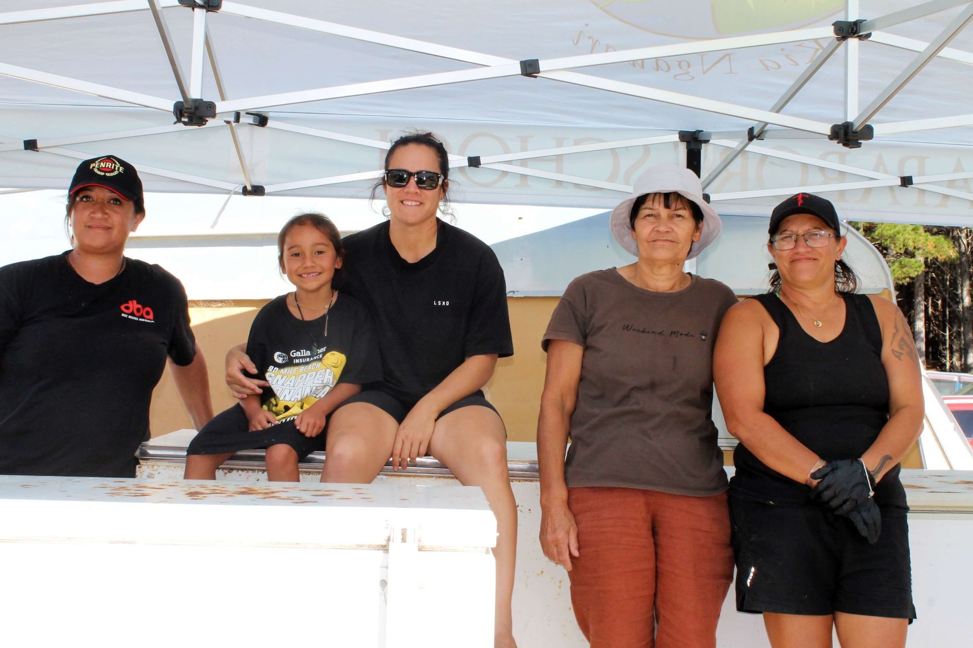 Five people, including a child, pose behind a white counter under a tent; sunny outdoor setting.