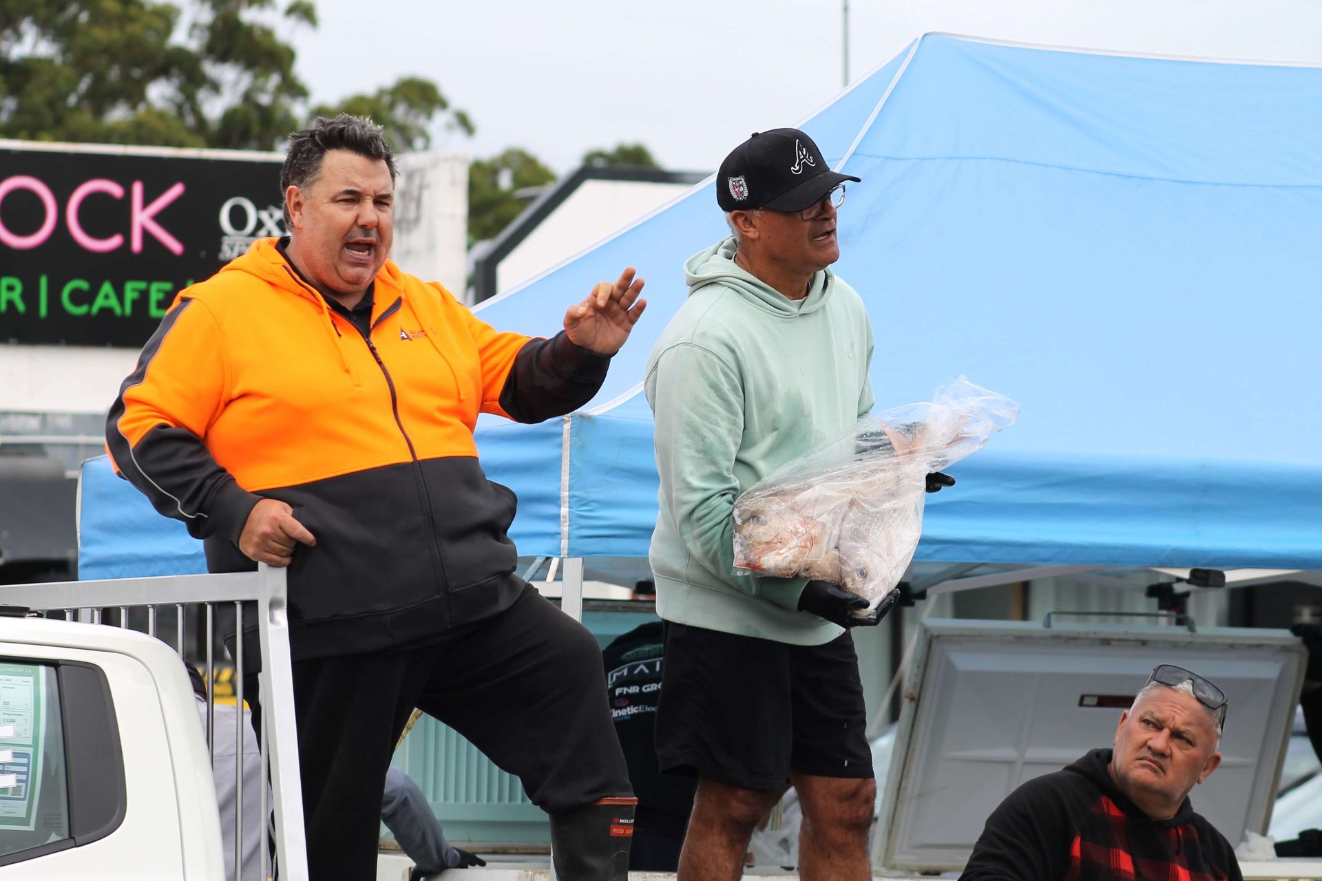 Man in orange jacket gestures, another carries a package, others observe near a tent.