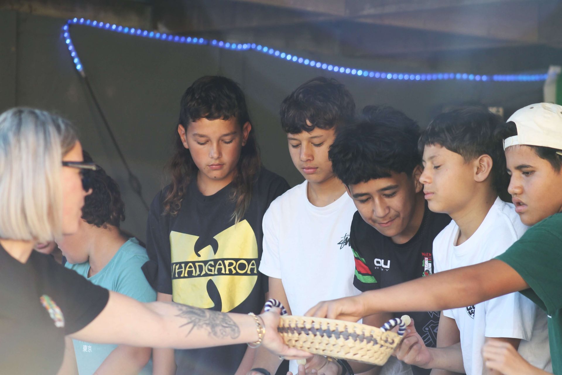 Woman offering food from a basket to a group of boys outdoors, string lights above.