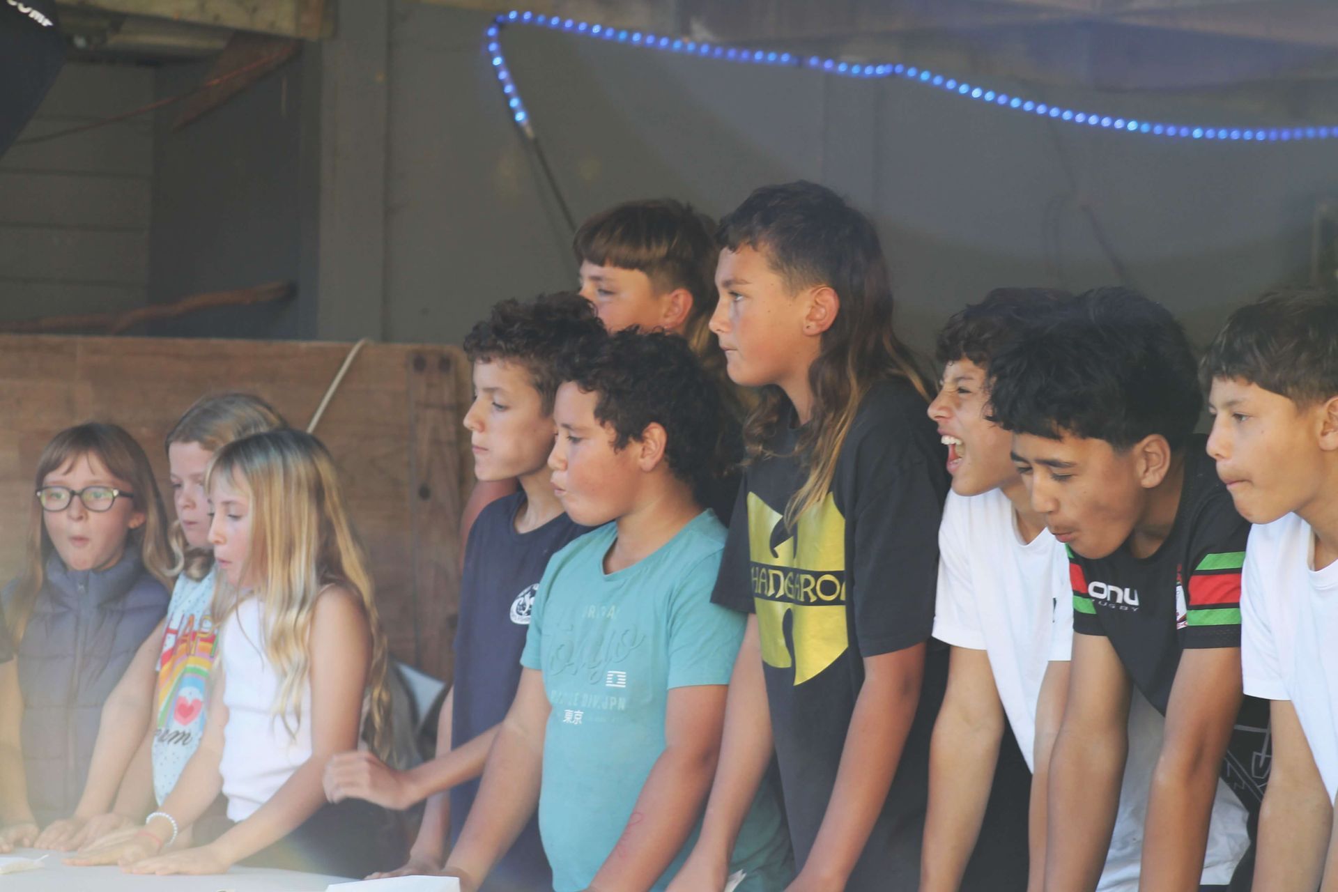 Children watching something outdoors under a wooden structure. They are smiling, and looking intently.