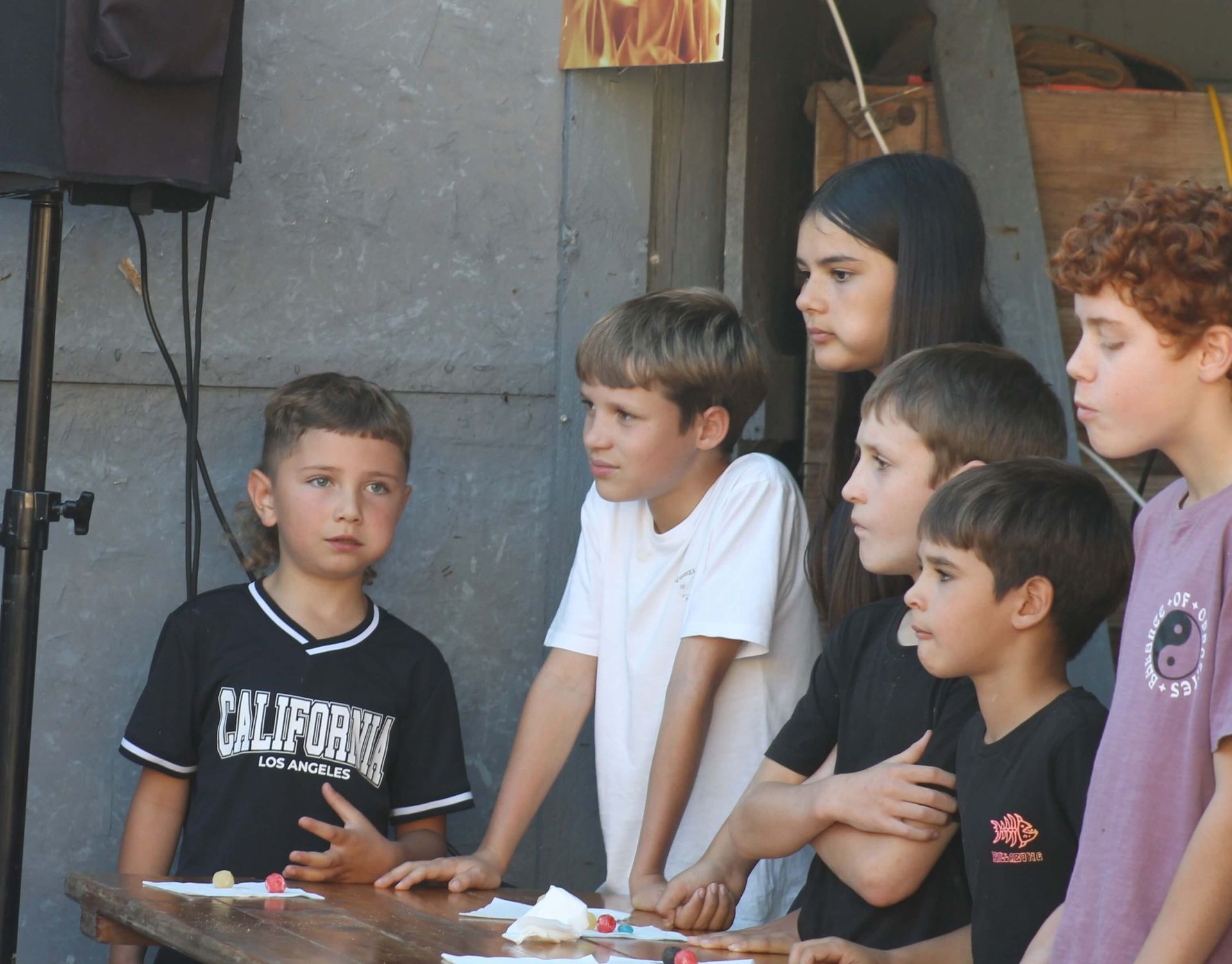 Children and a young woman gathered around a table; some looking at something, others with concerned expressions. Outdoors setting.