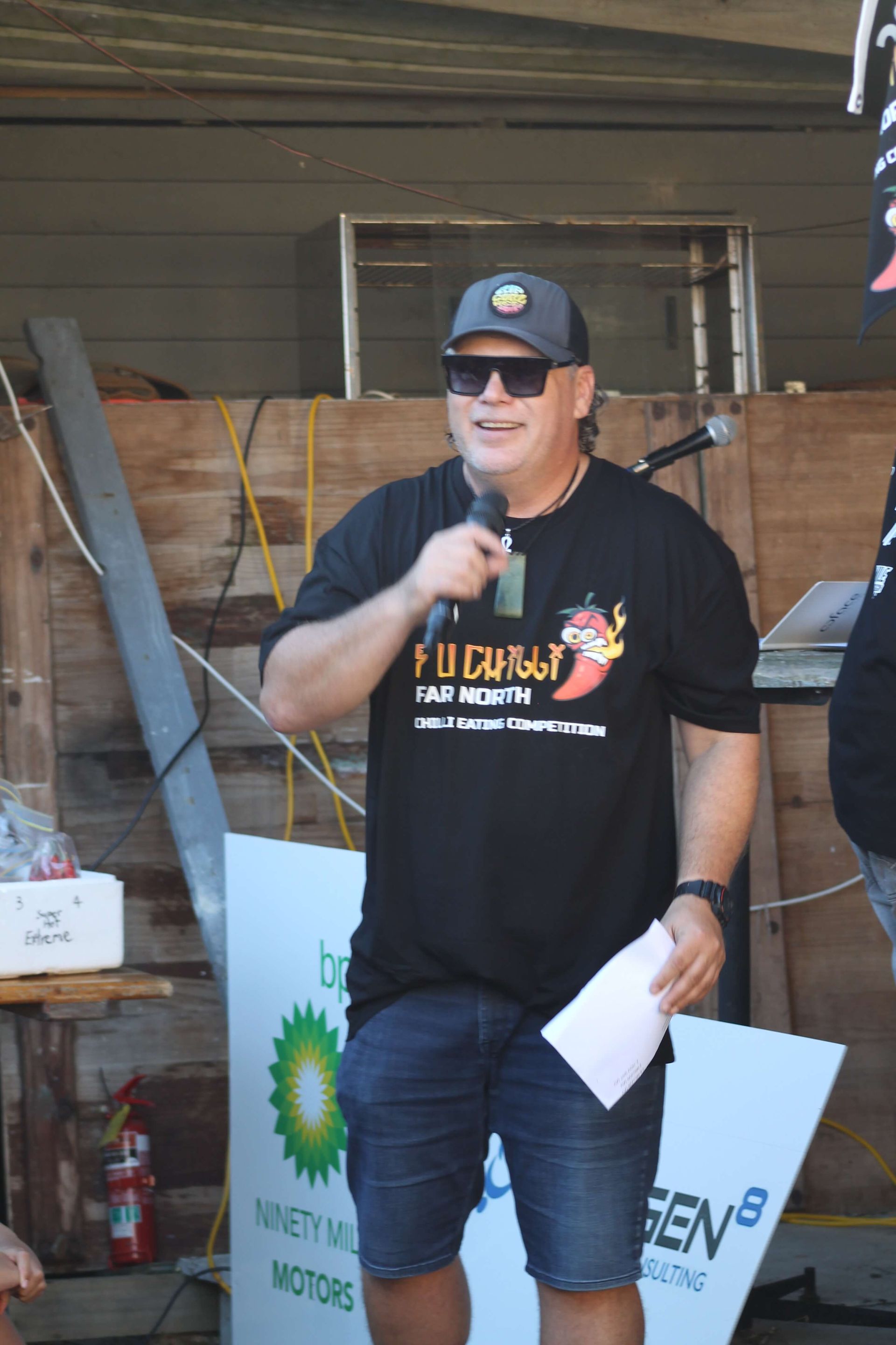 Man speaking into a microphone, wearing sunglasses, a hat and a t-shirt at an outdoor event.