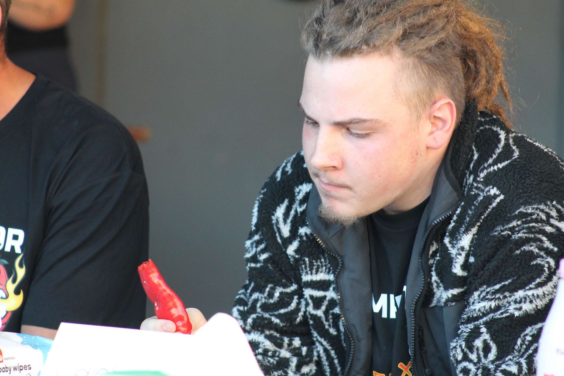Man with dreadlocks looking down, holding a red object. Wearing a patterned jacket and black shirt.