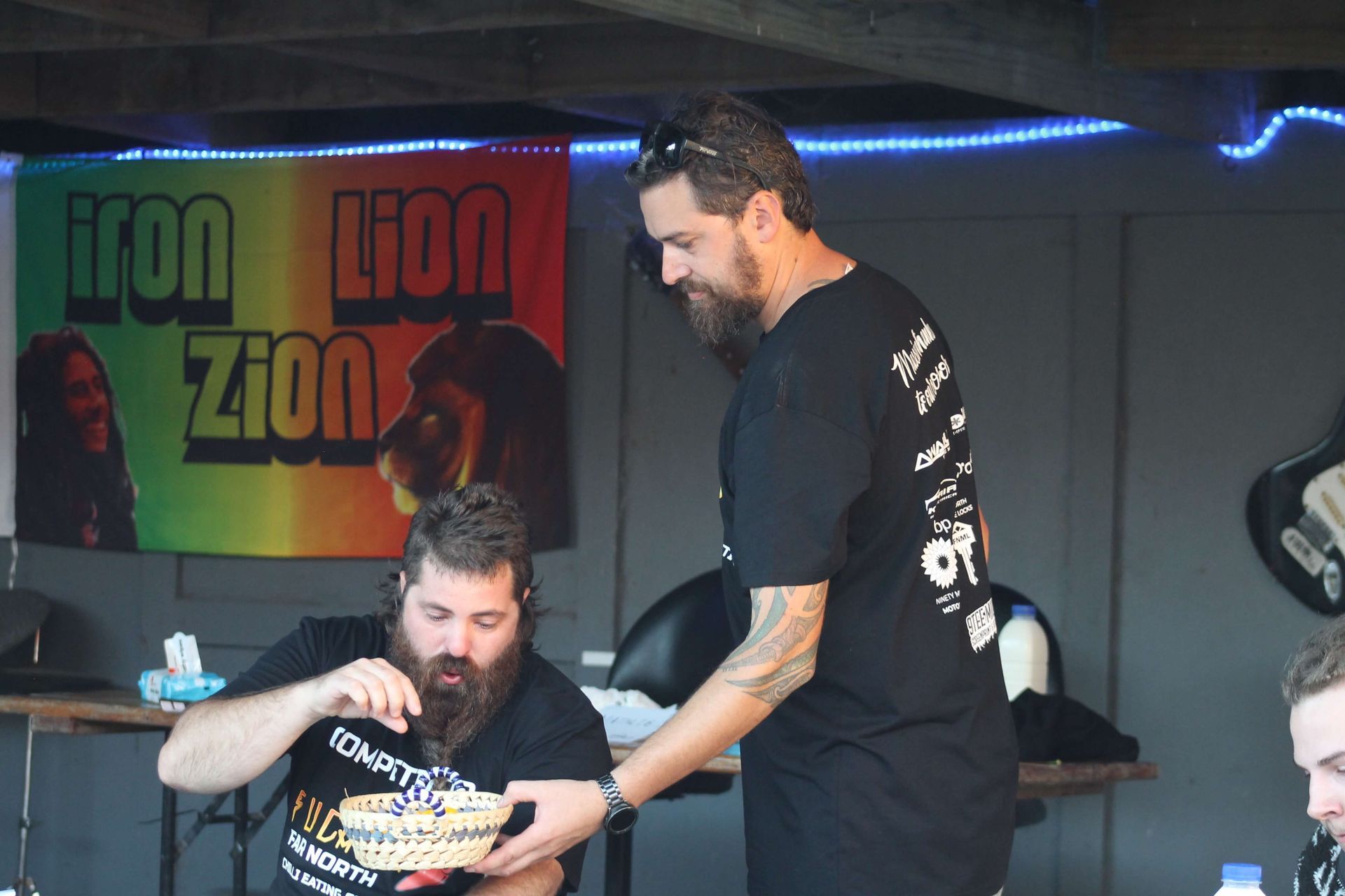 Two bearded men at a table. One looks into a bowl. The other reaches toward it.