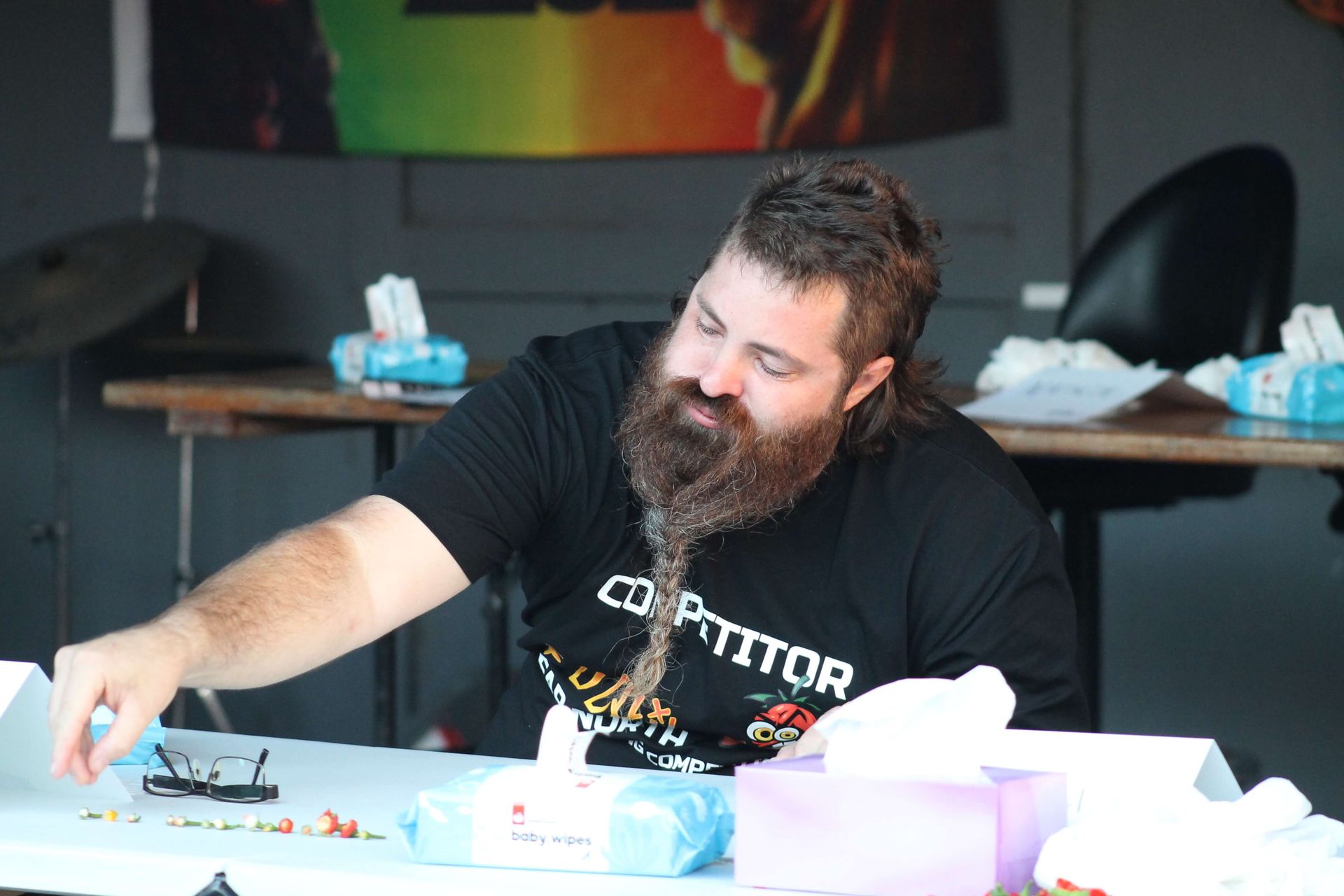 Man with braided beard sorting items on a table with tissues, in front of a flag.