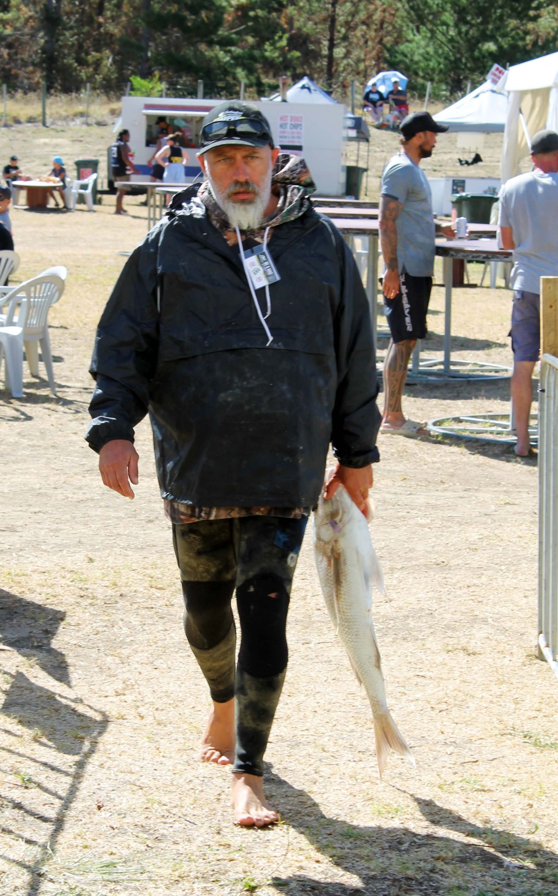 Man holding a fish, walking barefoot on gravel. He wears a jacket, leggings, and a cap. People and tables in background.