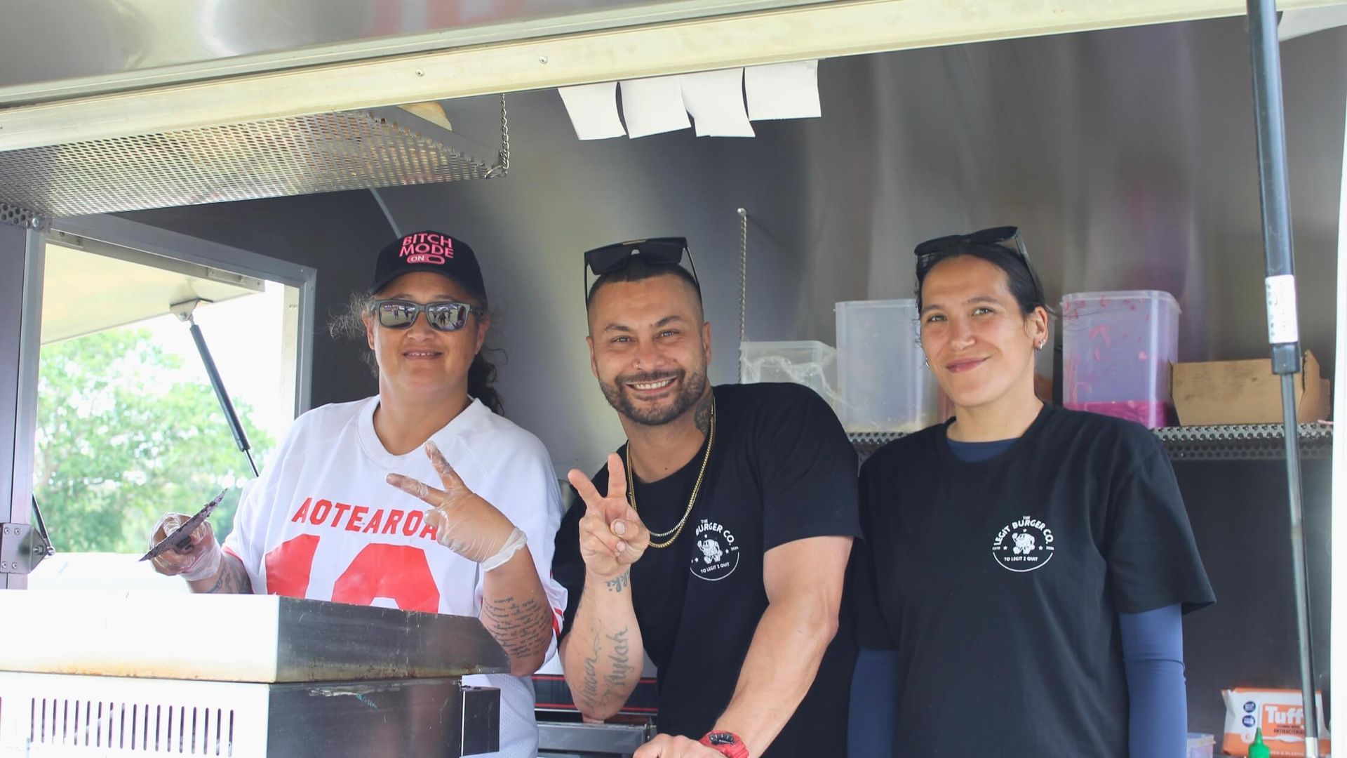 Three people smiling in front of a food truck. Man gives peace sign. Other two wear matching t-shirts.