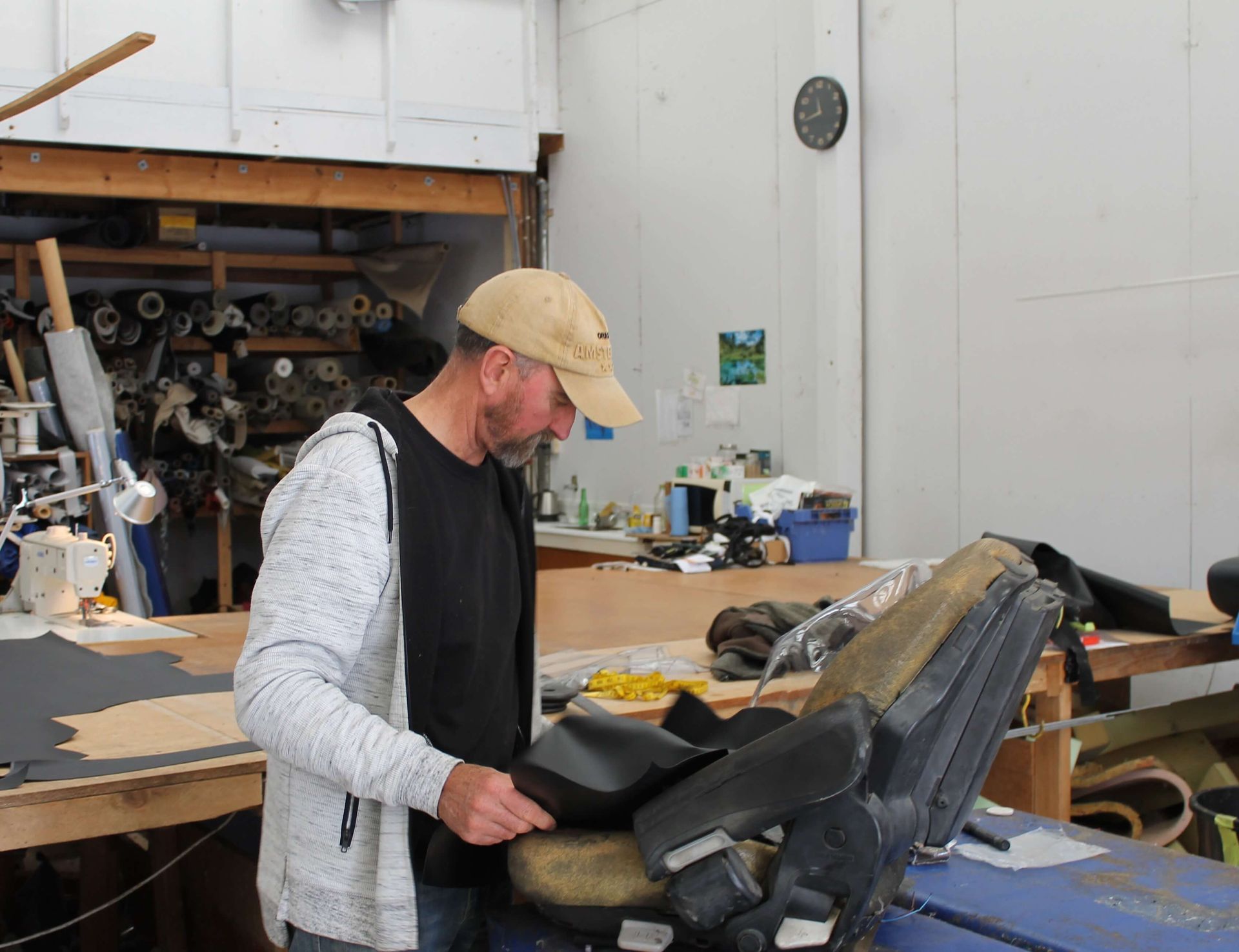 Man in a workshop inspecting black fabric on a car seat; rolls of fabric in the background.