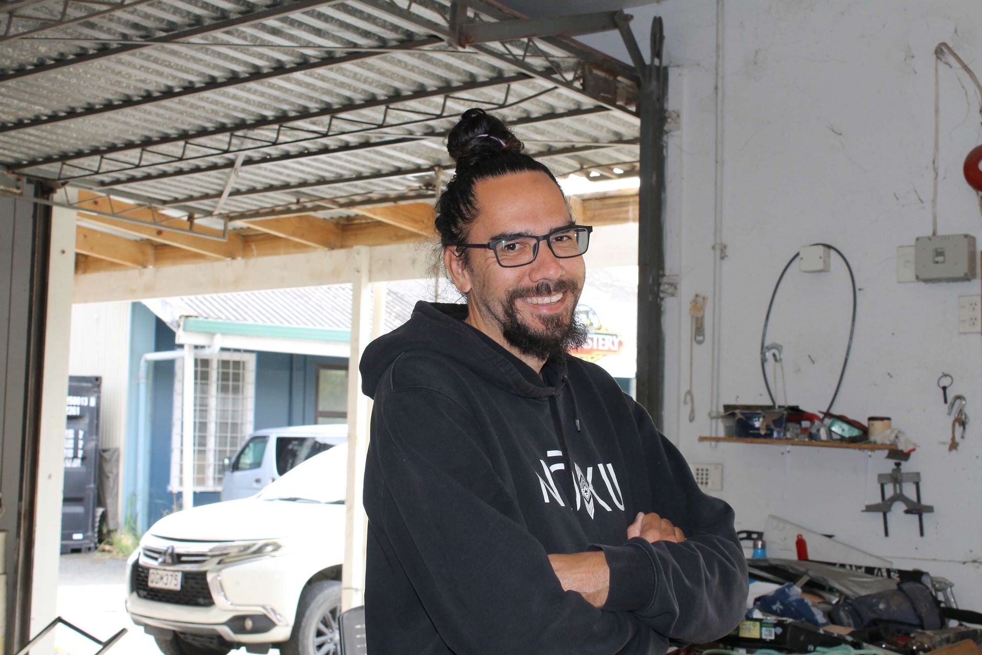 Man with glasses and bun smiling in garage; car and tools in the background.