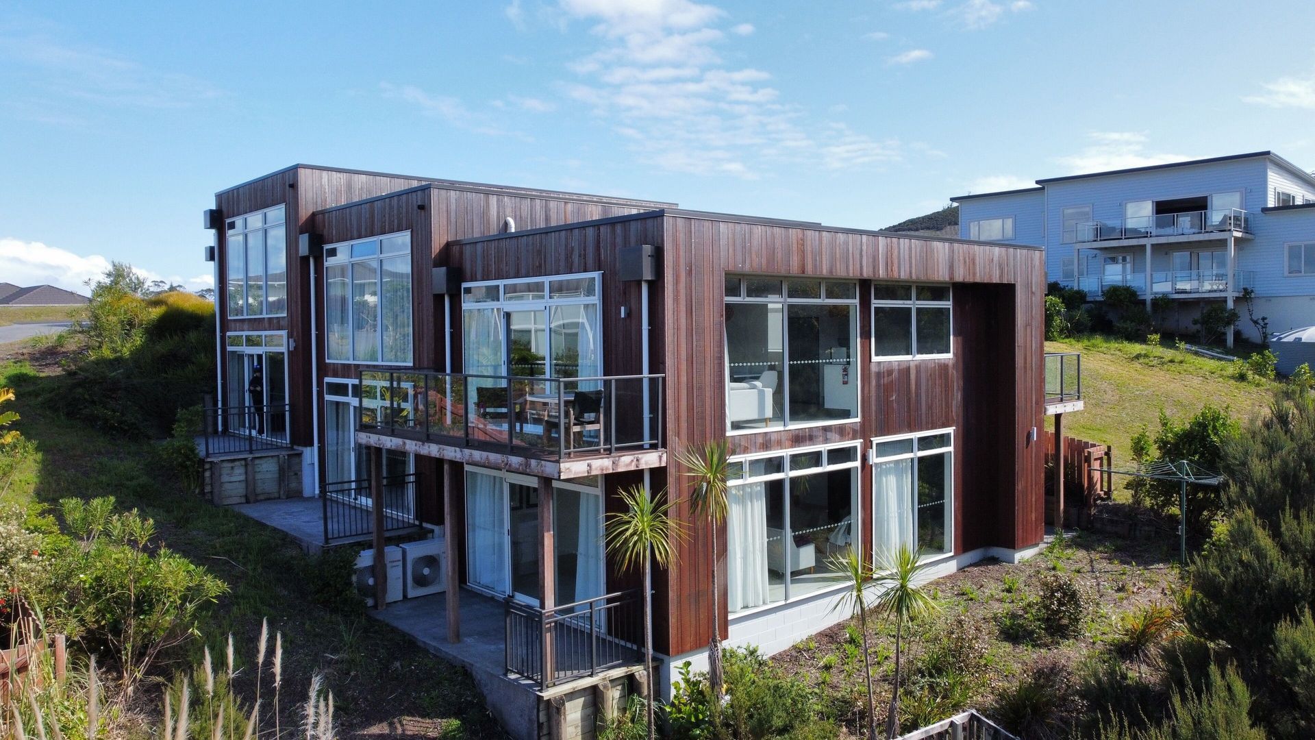 Modern two-story house with dark brown siding and large windows on a sunny hillside.