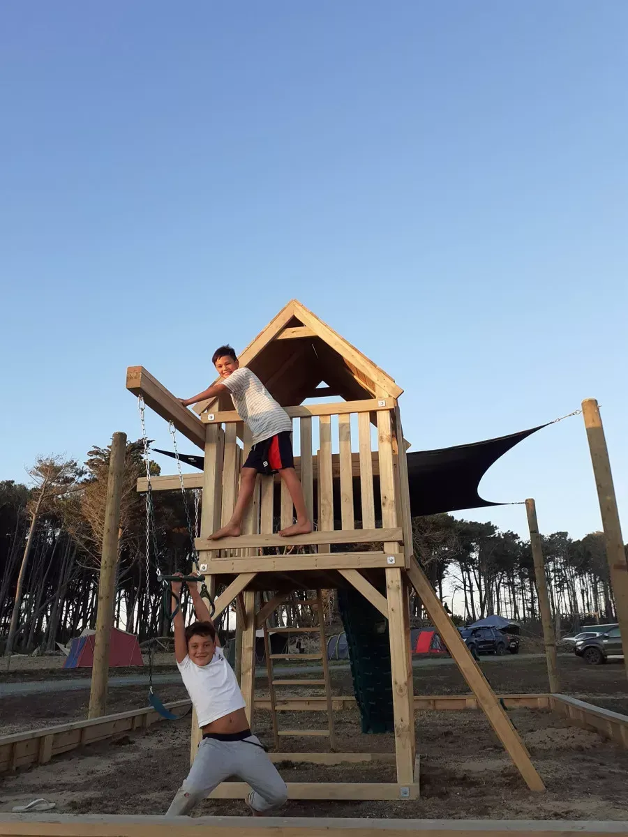 Two people playing on a wooden playground structure outside. One is standing on the top level, one hanging from a bar below.