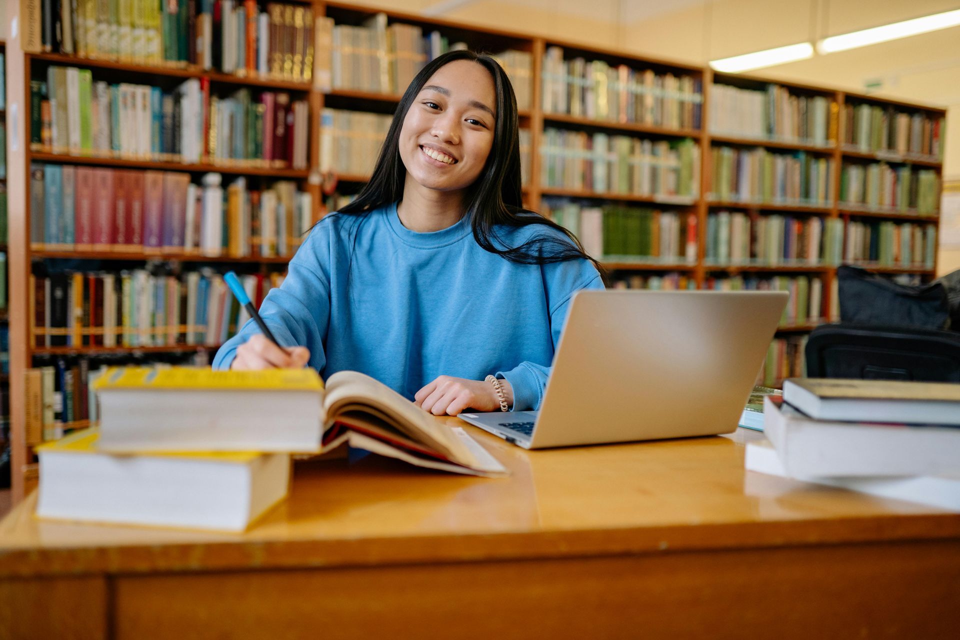 Woman with laptop, books, and pen smiles in a library.