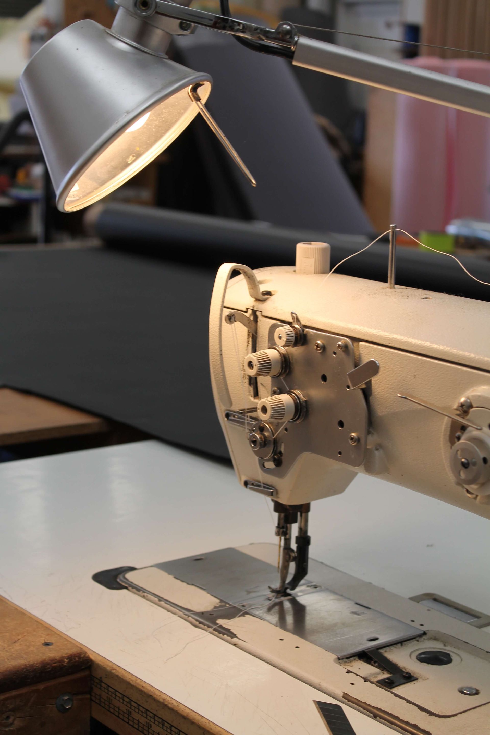 A lit industrial sewing machine with thread, on a white table. Light illuminates the metal needle and base.