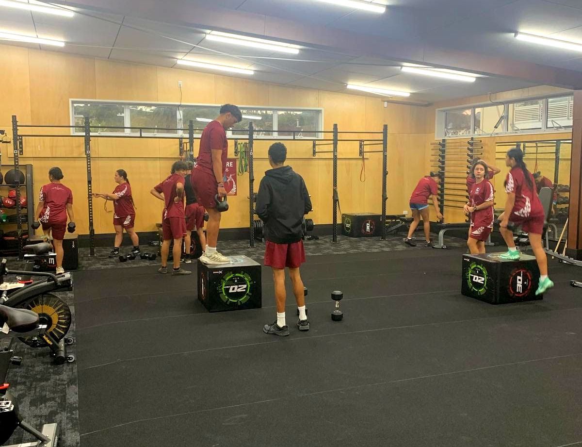 People in maroon athletic wear exercising in a gym. Jumping on boxes, weights, and pull-up bars are visible.