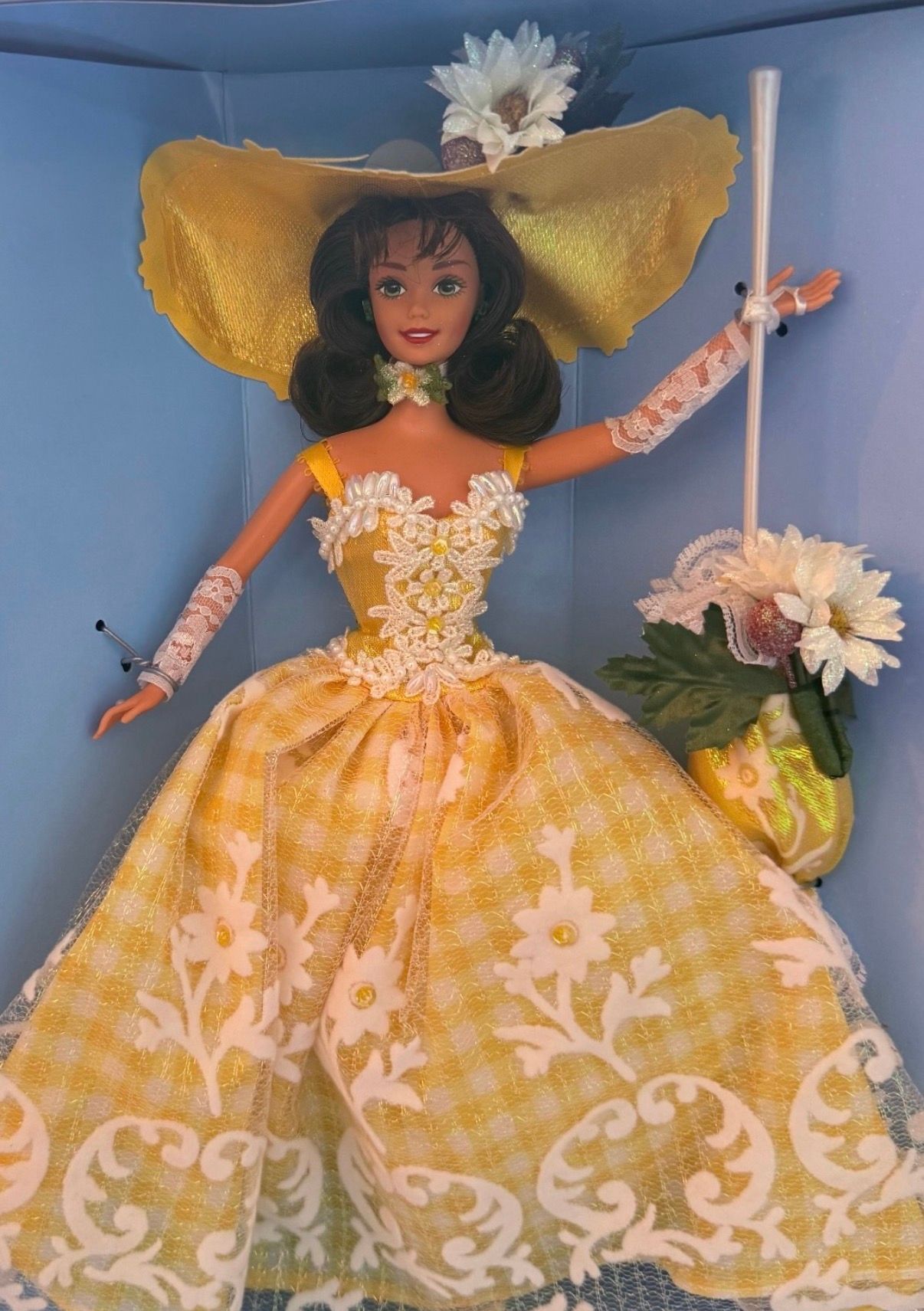 Doll in a yellow floral dress and wide hat, posed in a blue display box with flowers.