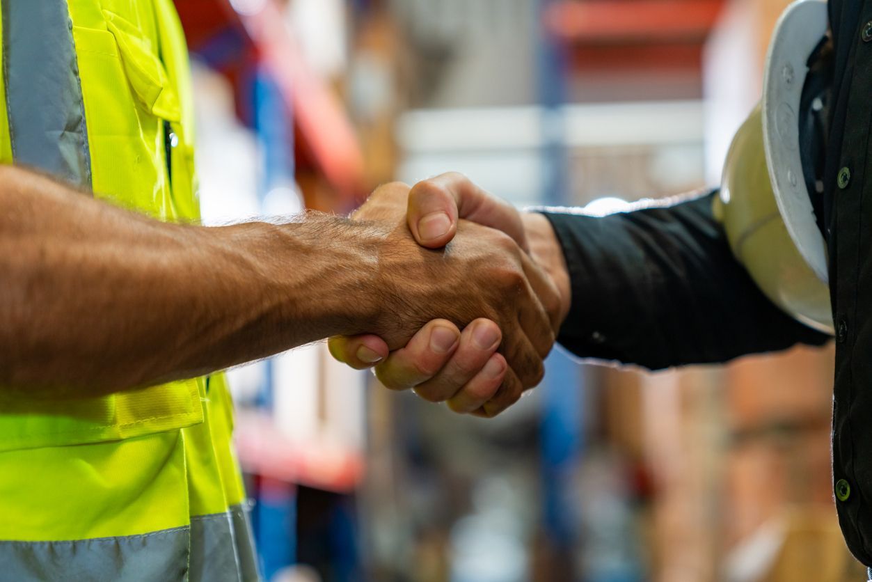 Two workers in a warehouse shake hands, one wearing a safety vest and the other holding a hard hat.
