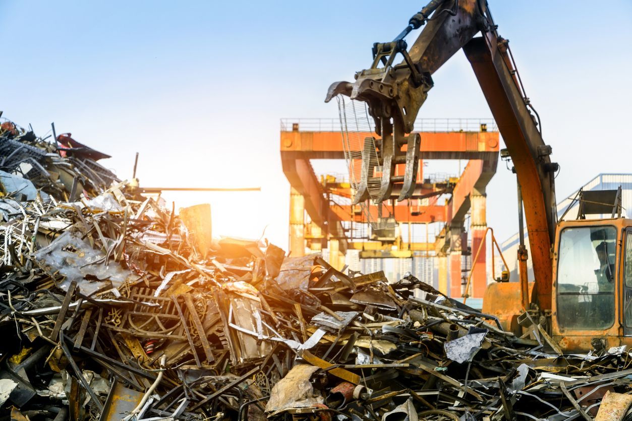 An excavator with claw arm picks up scrap metal in a junkyard. Bright sunlight, orange machinery.