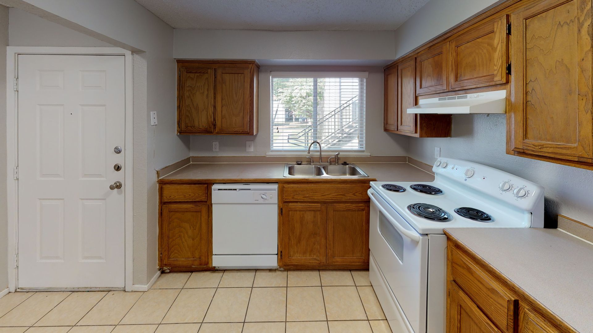 Empty kitchen with white appliances, wood cabinets, tiled floor, and a window over the sink.