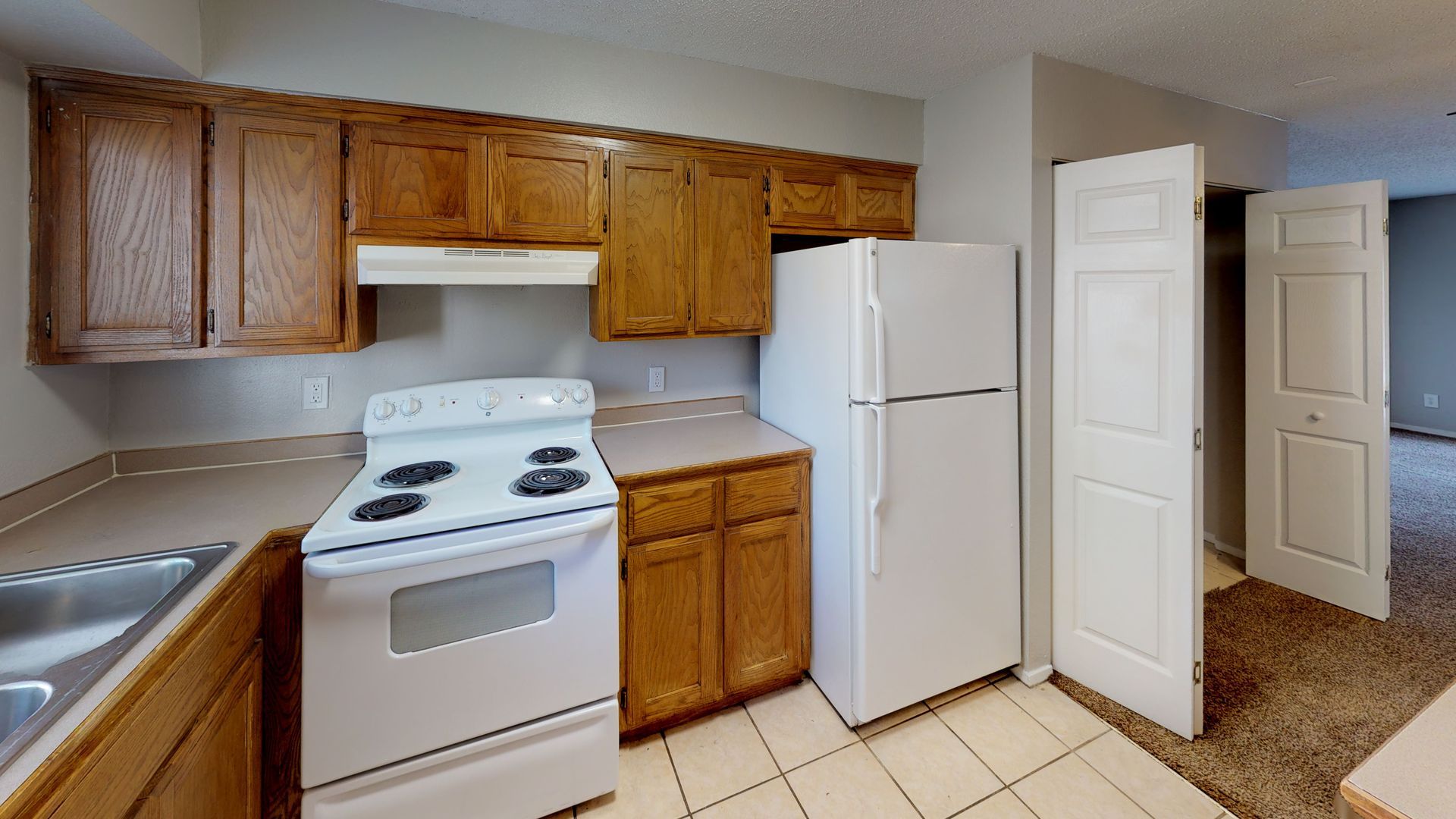 Small kitchen with wood cabinets, white stove and refrigerator, tiled floor, and open door to carpeted area