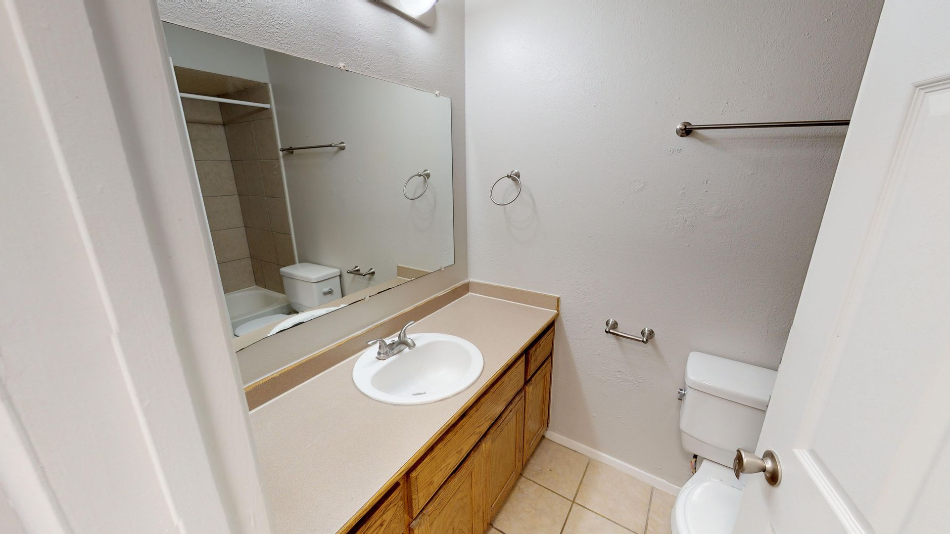 Small bathroom with a vanity sink, large mirror, toilet, towel bars, and beige tile floor.