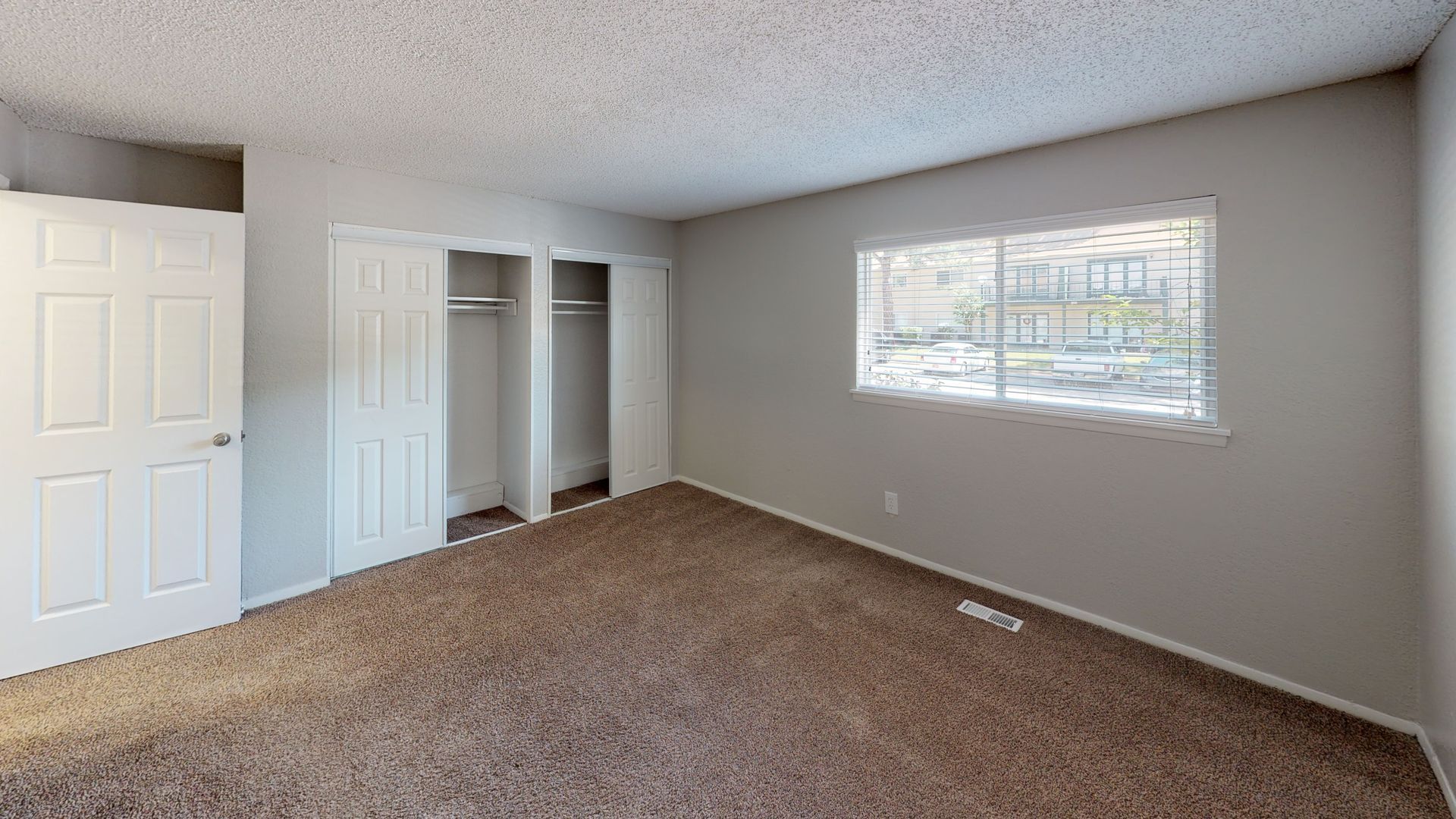 Empty carpeted bedroom with gray walls, white closets, and a window with blinds