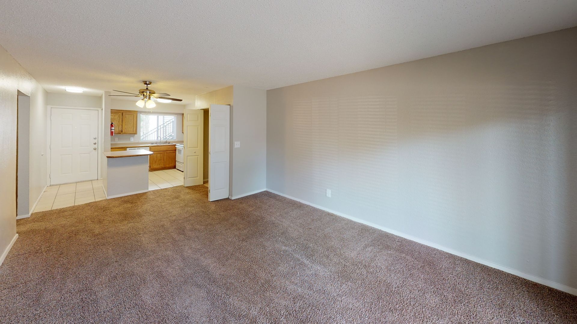 Empty carpeted living room with beige walls and a view into the kitchen and hallway