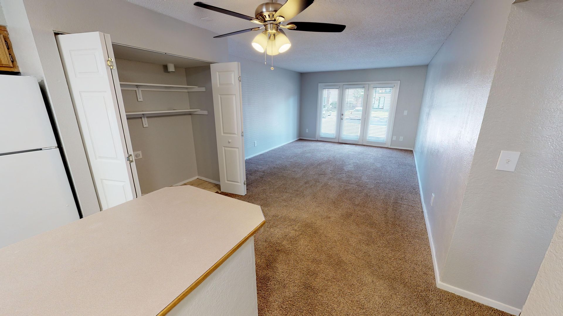 Empty carpeted room with ceiling fan, closet shelving, kitchen counter, and French doors to a patio.
