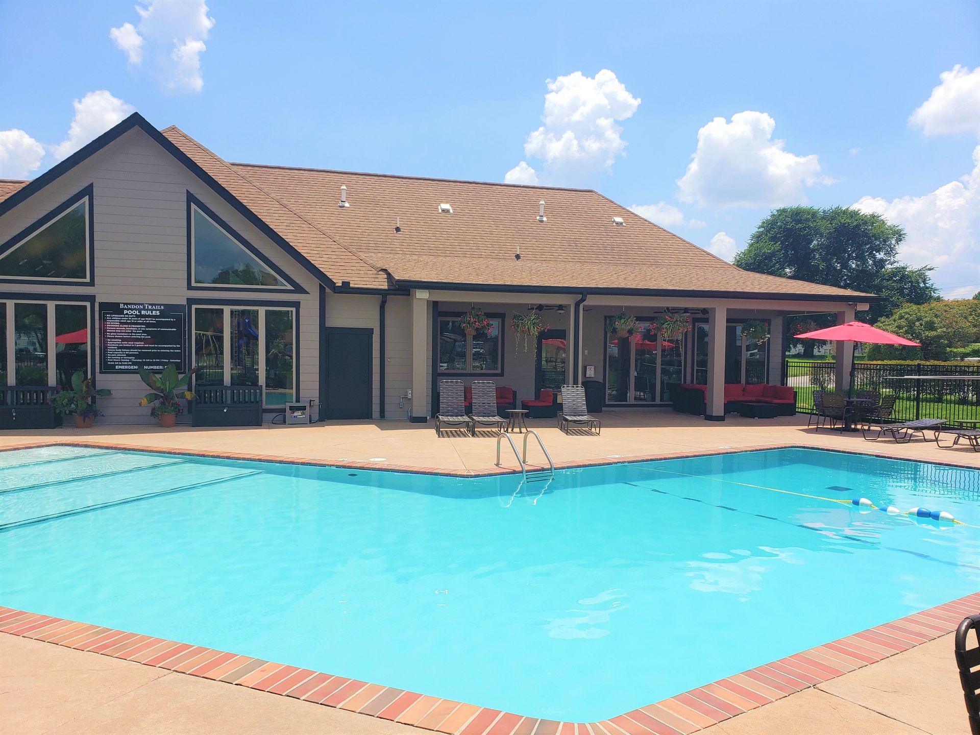 Outdoor swimming pool beside a tan clubhouse with lounge chairs and red umbrellas under a blue sky