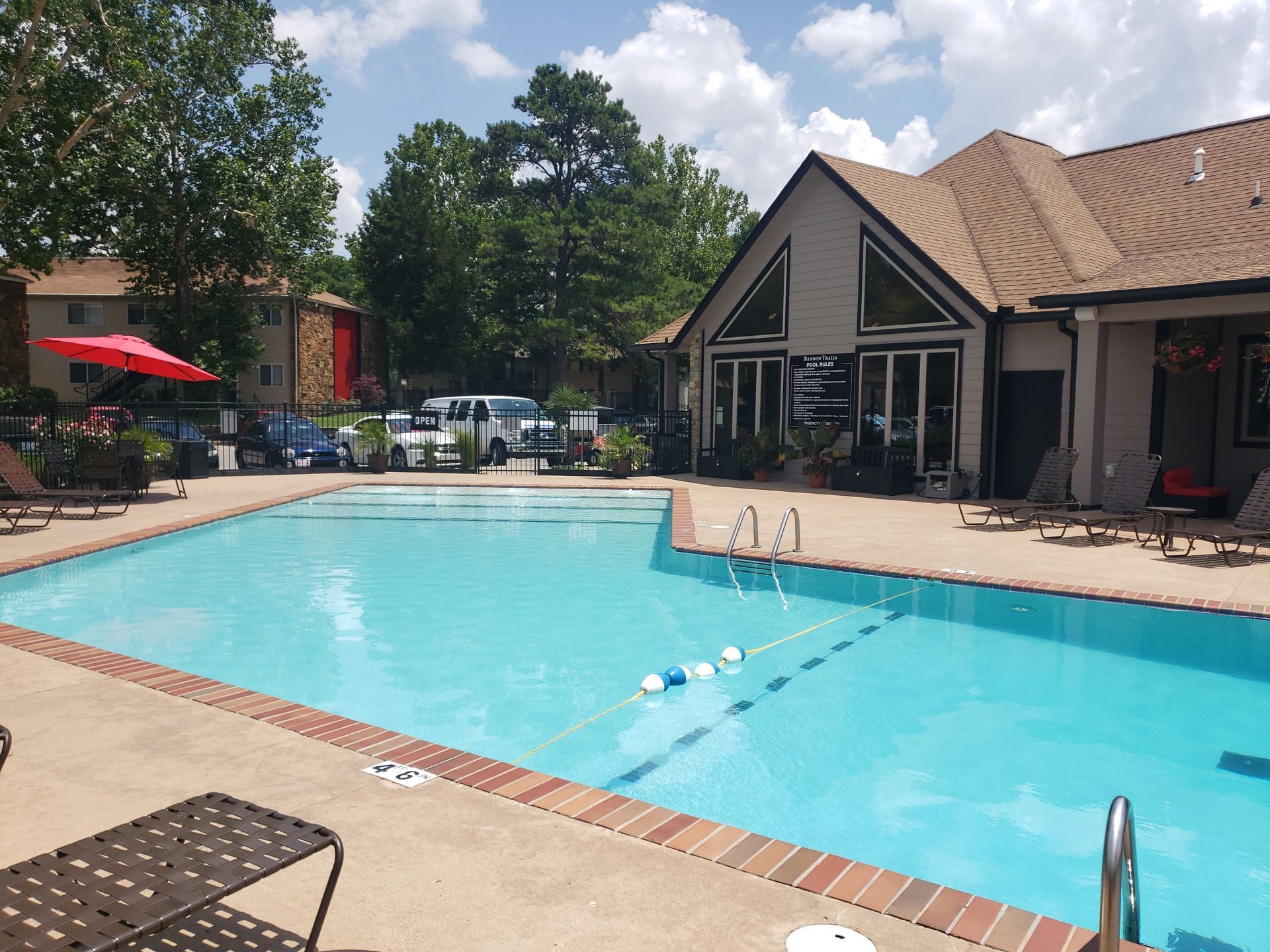 Outdoor swimming pool beside a beige building with lounge chairs, trees, and red umbrellas on a sunny day
