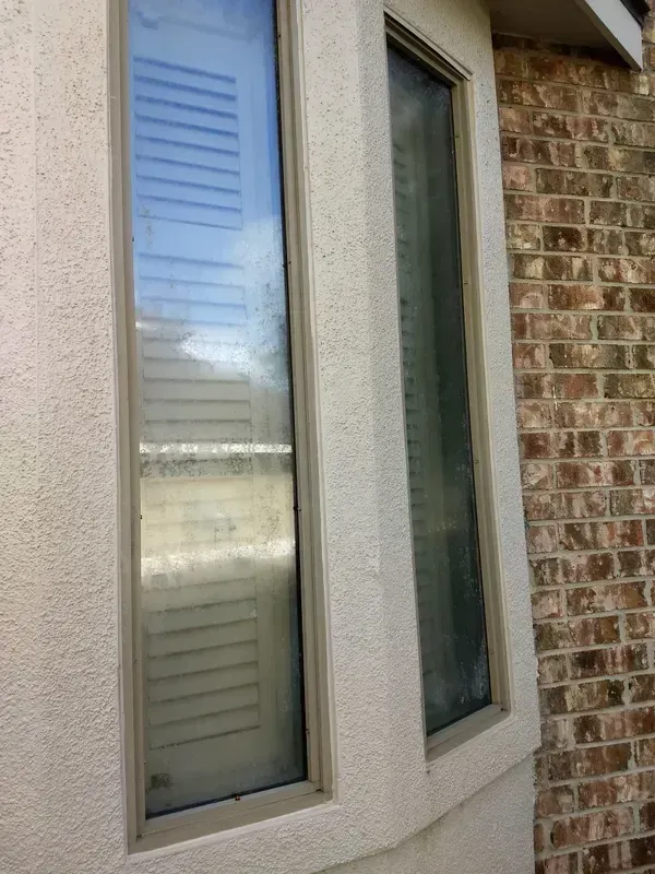 Two tall, narrow windows in stucco wall, beside brick wall. One window has interior blinds.