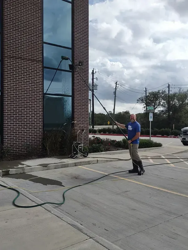 Man washing building windows with a long-handled tool; outdoors, cloudy day, brick building.