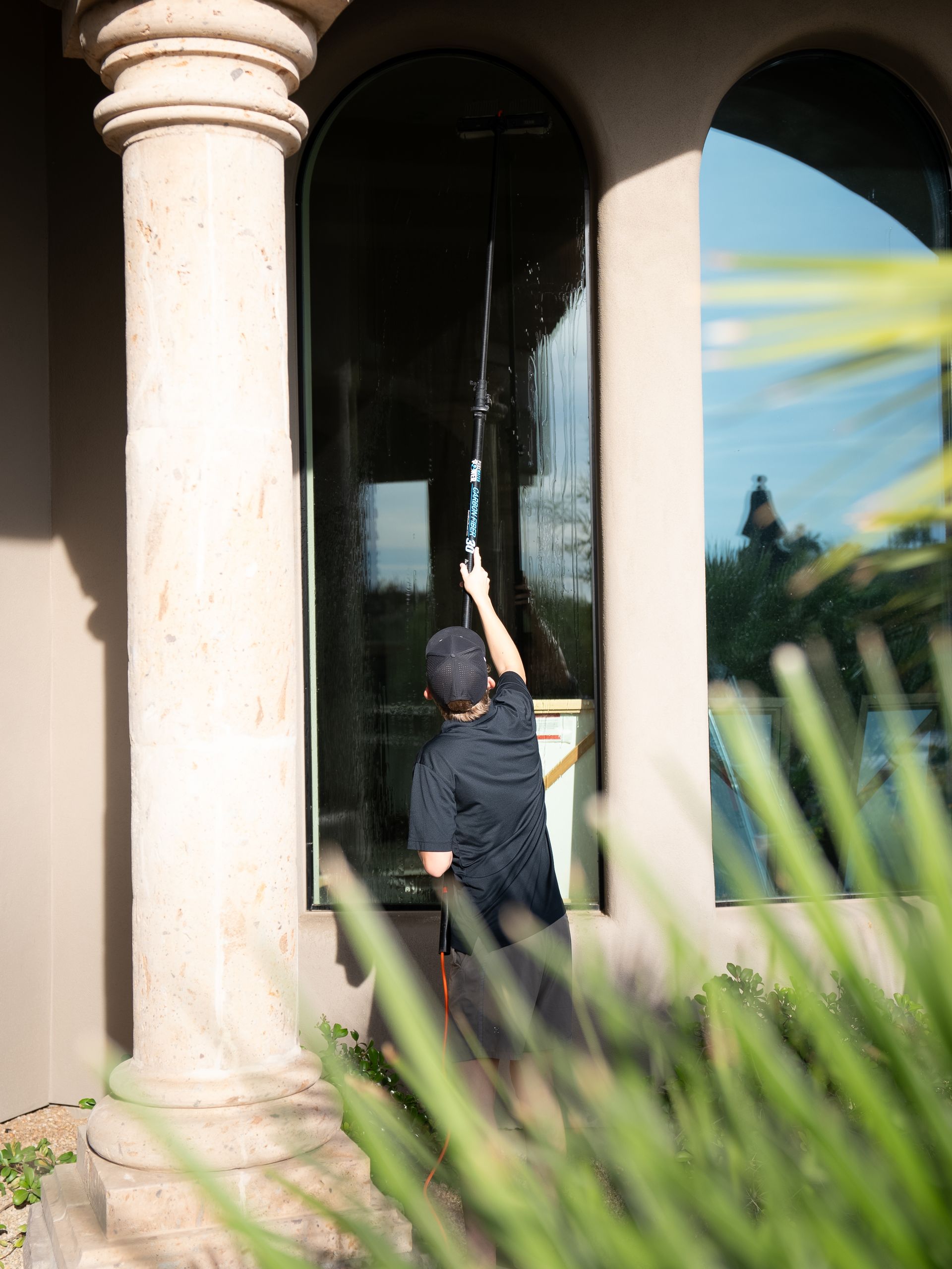 Person reaching up to touch a dark arched window beside a stone column outdoors.