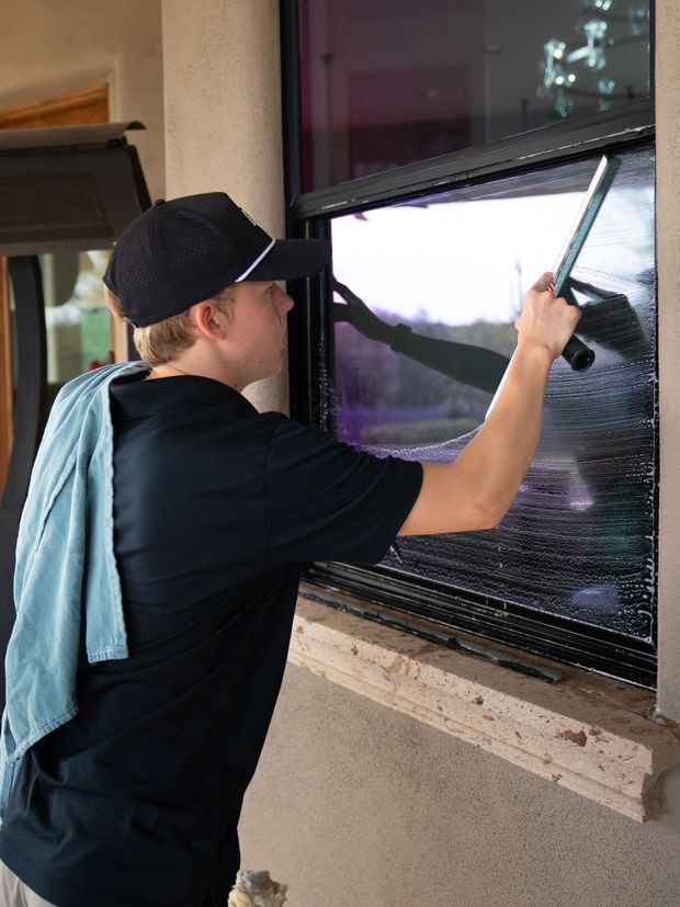 Person cleaning a window with a squeegee, wearing a black cap and shirt, outdoors on a porch.