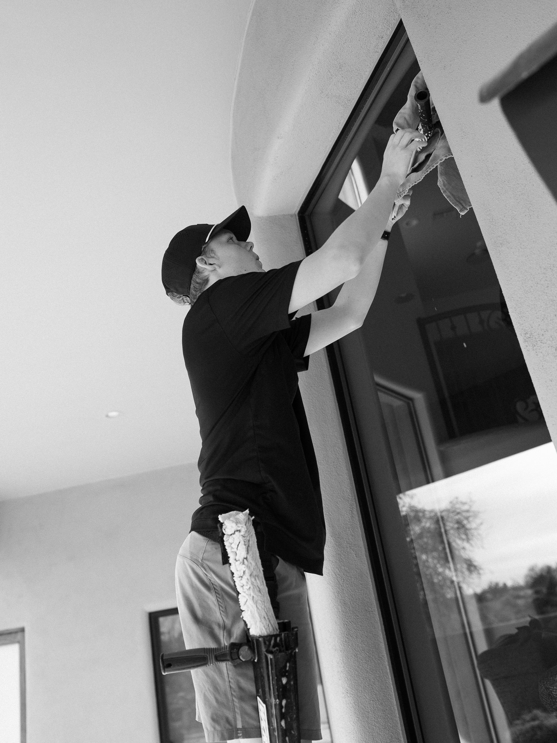Person standing on a ladder, reaching up to work on a window frame indoors.
