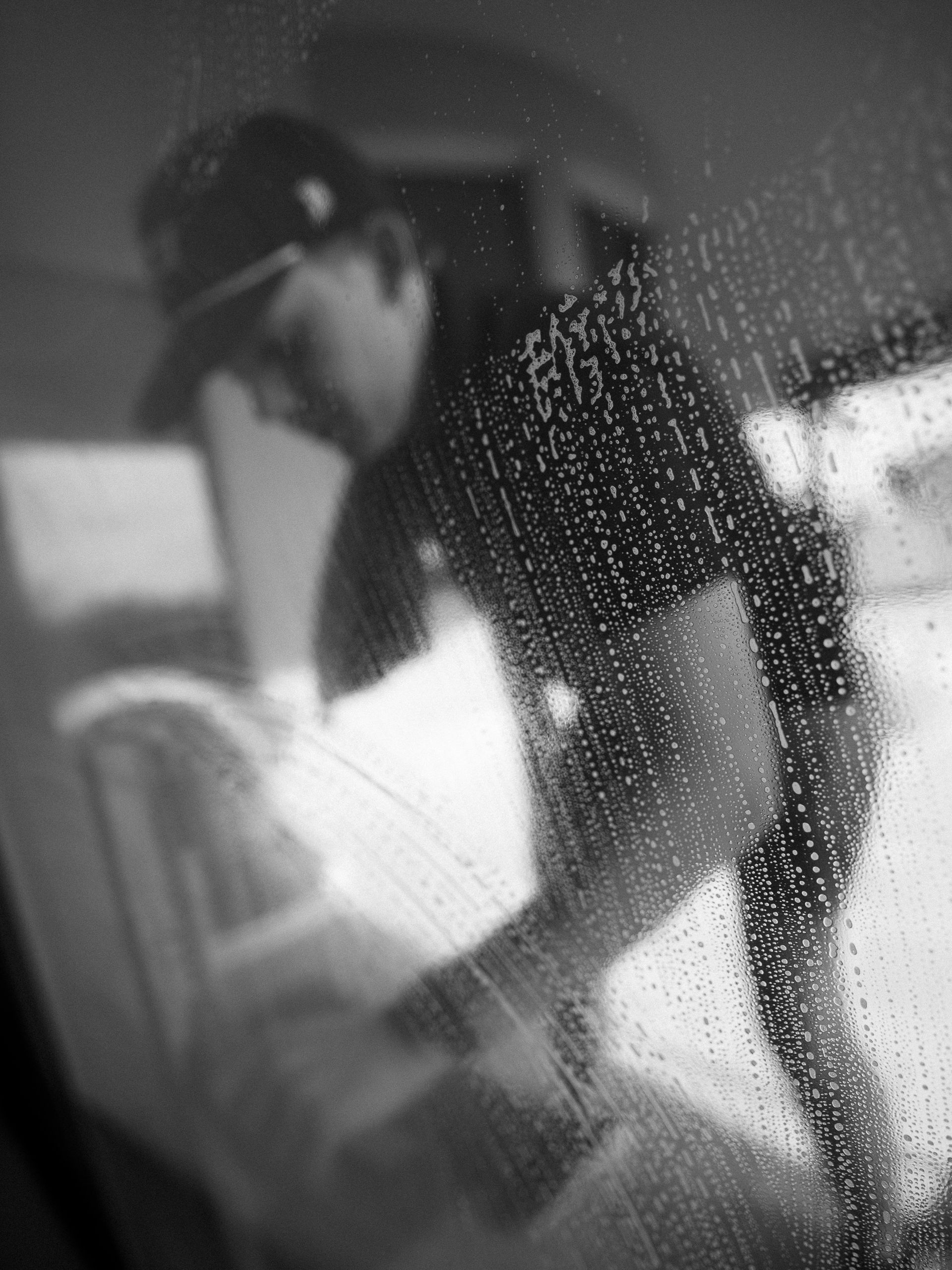 Person in dark clothing and cap leans over a bed in a dim room, seen through a rain-speckled window.