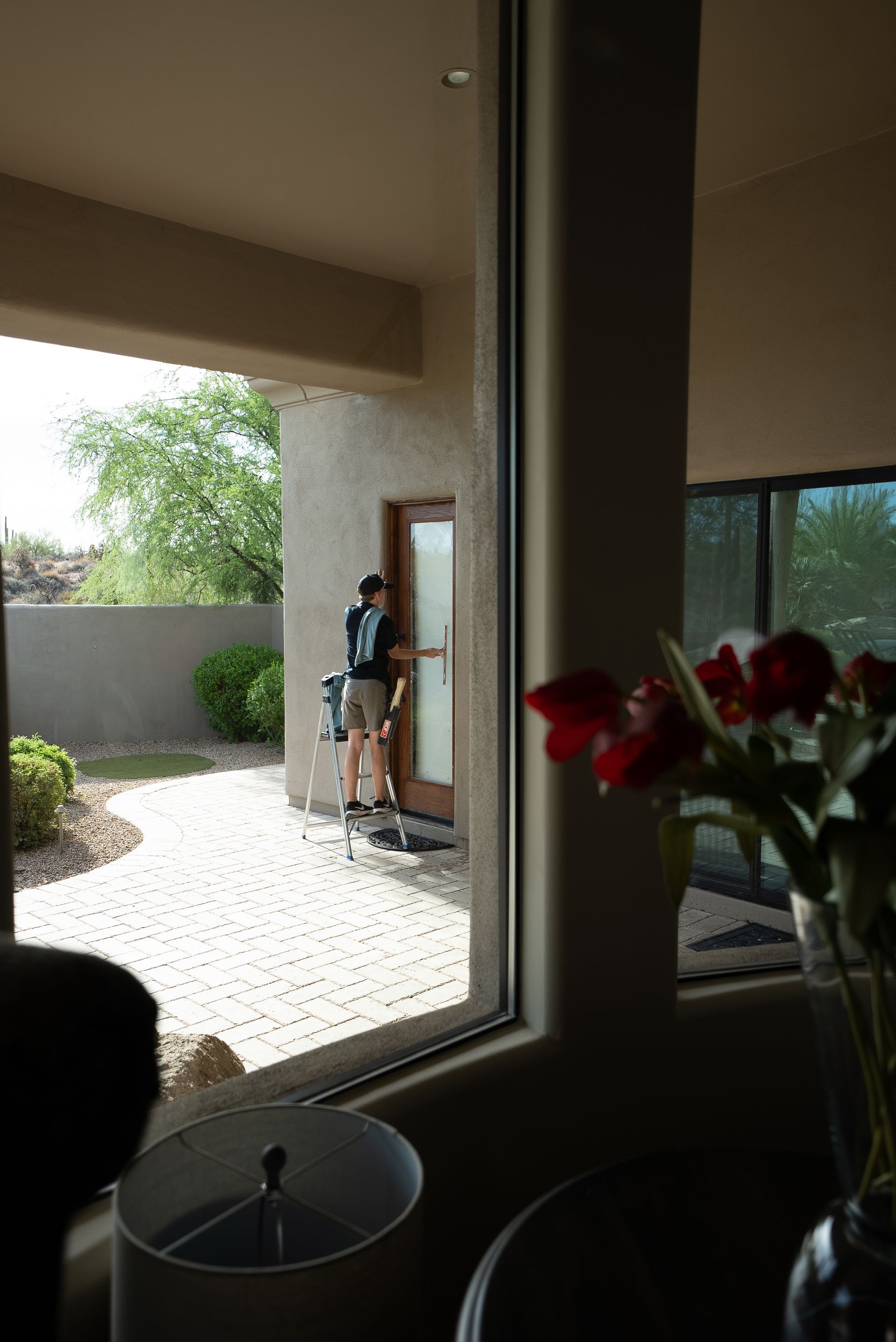 Person standing by a doorway in a sunlit courtyard, seen through an indoor window.