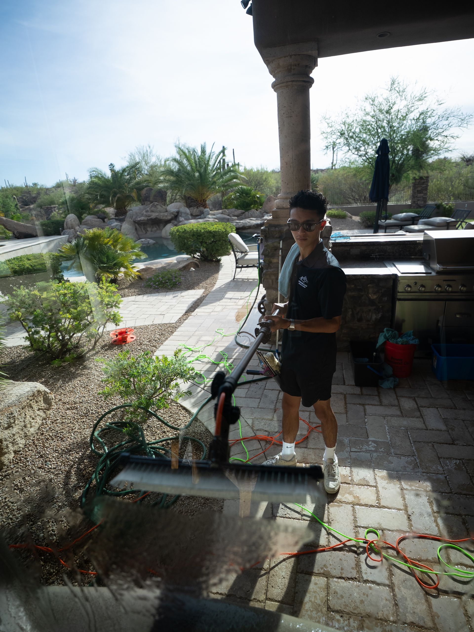 Person using a pressure washer in a garden patio area, spraying water near plants and a stone walkway.