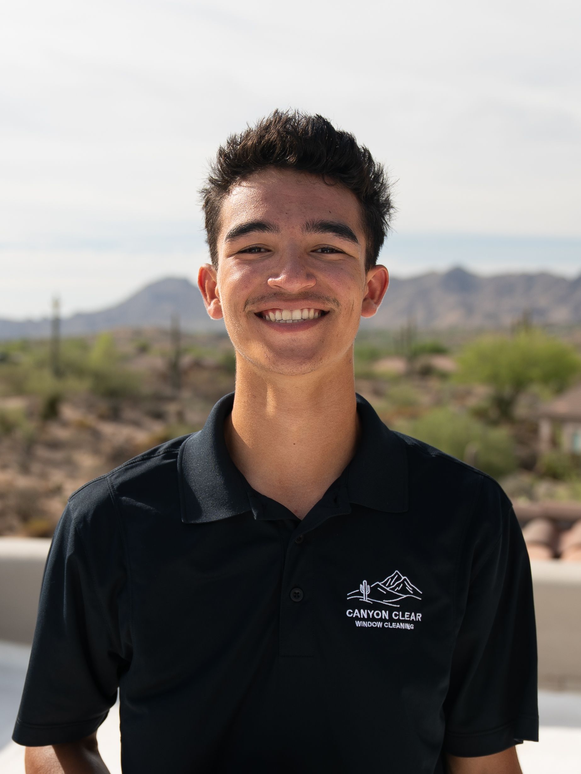 Smiling person in a black polo shirt standing outdoors with mountains in the background