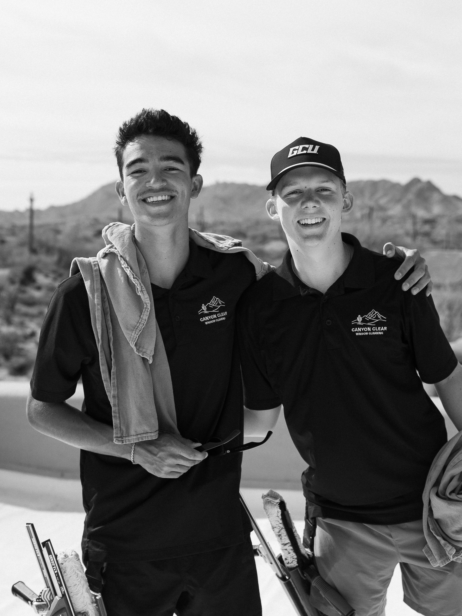 Two people in black shirts smiling outdoors, one holding a towel, with mountains in the background.