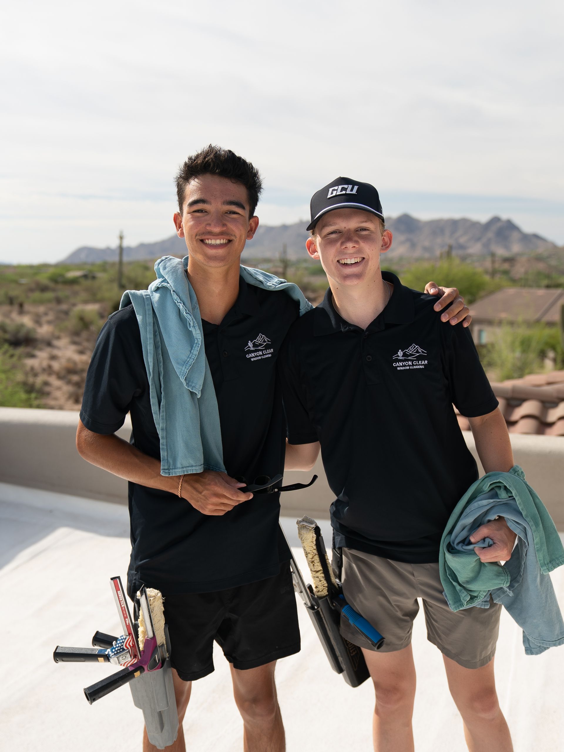 Two smiling hikers in black shirts posing with towels, desert mountains in the background.