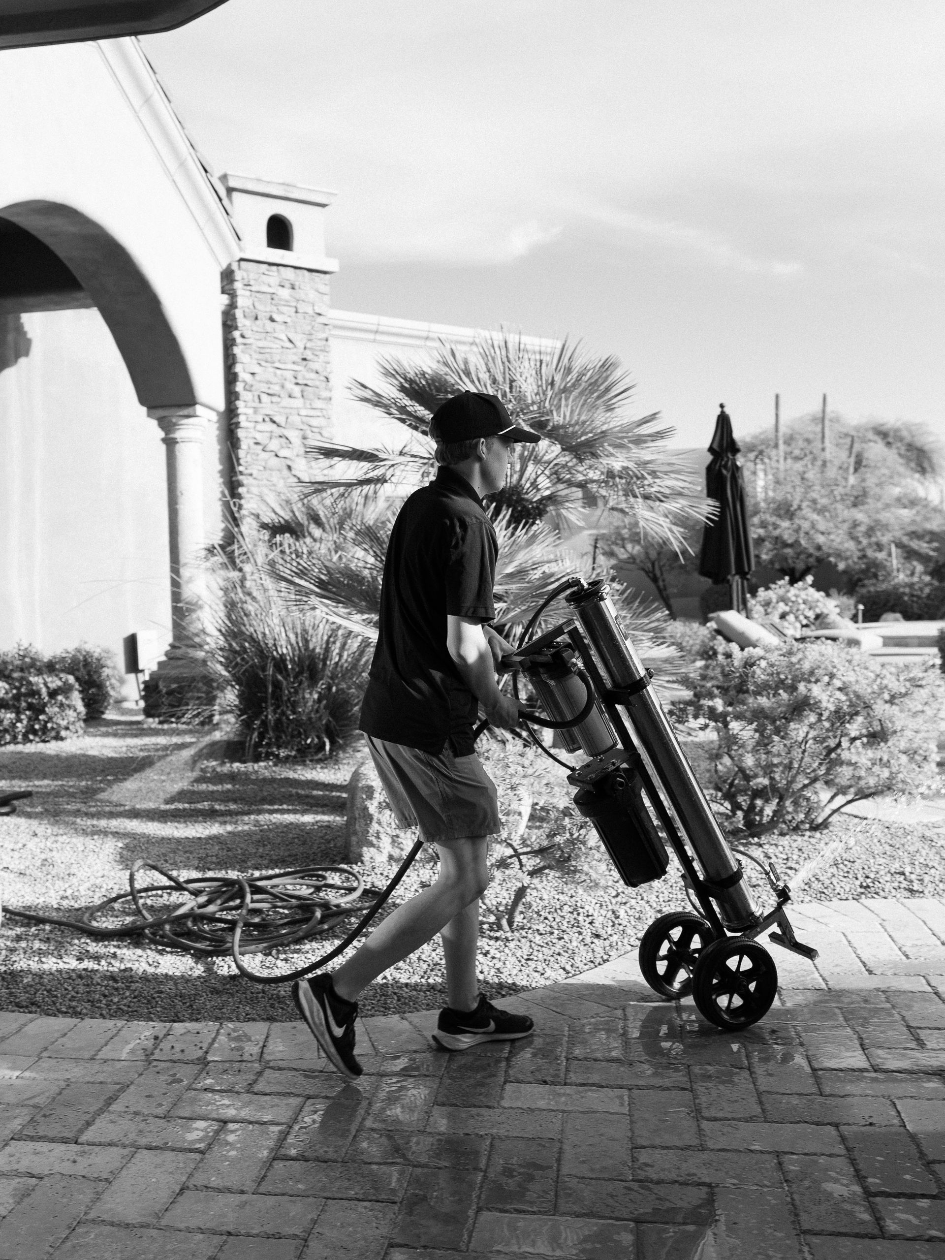 Person pushing a golf bag on a paved path beside a building and landscaping.