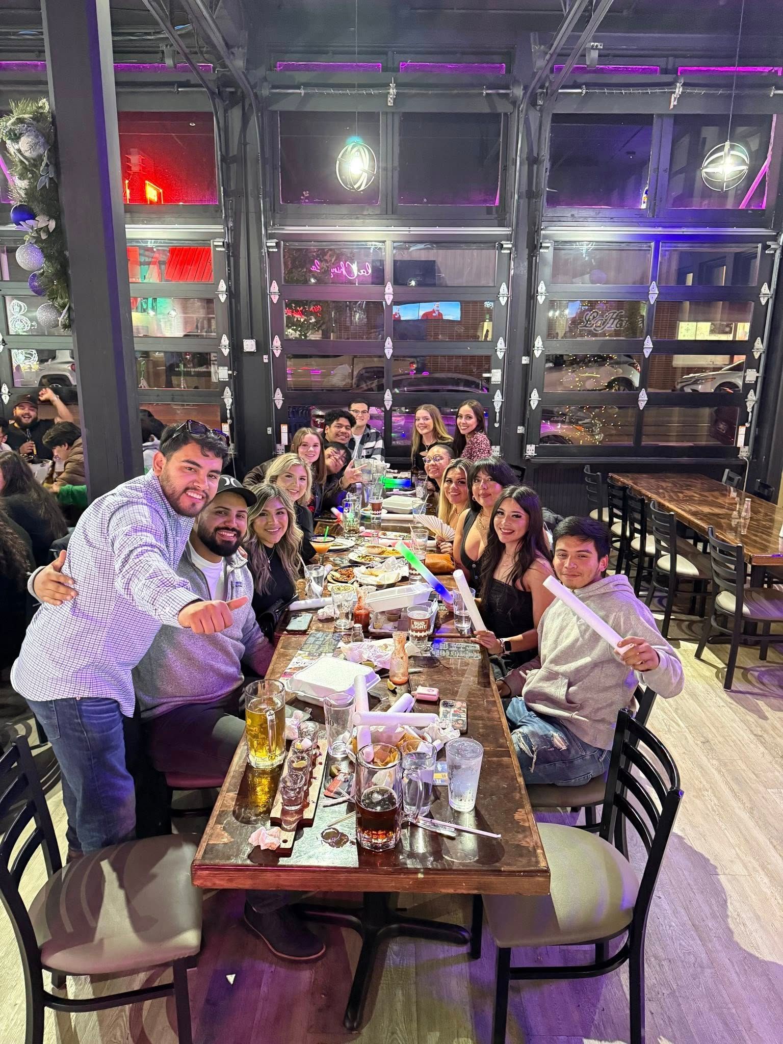 Group dining at a long restaurant table, with people smiling and raising drinks in a festive indoor setting
