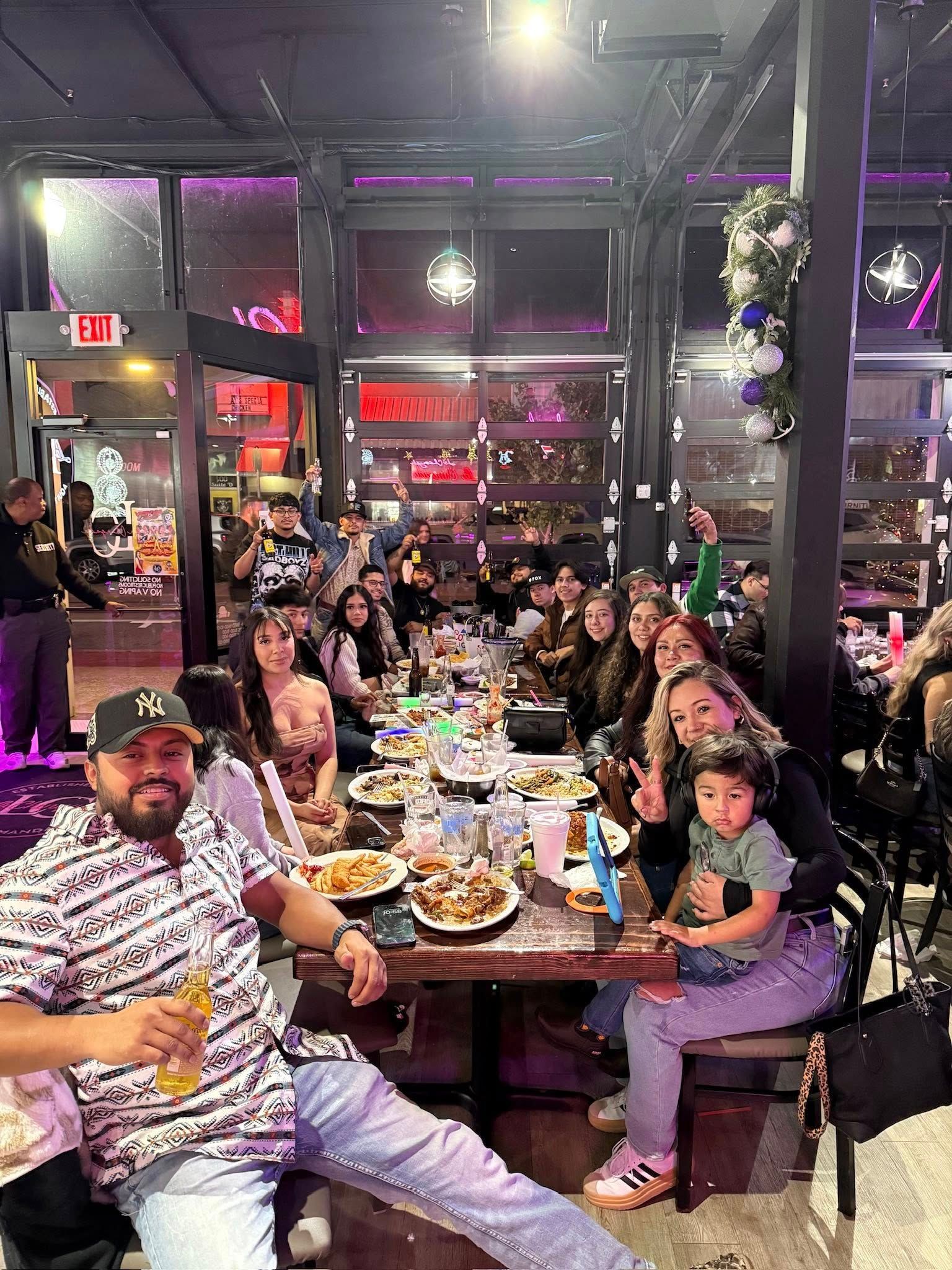 Large group dining at a restaurant, seated around a crowded table with plates, drinks, and smiling faces.