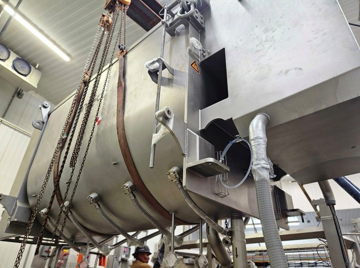 Large stainless steel industrial meat processing machine suspended by chains in a factory.