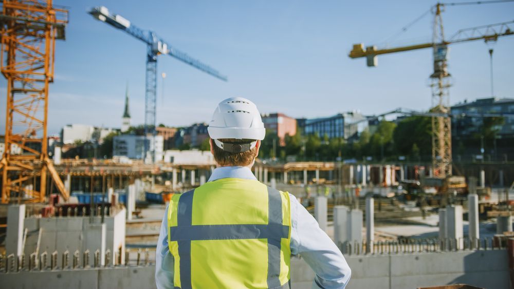 Construction worker in hard hat and vest surveying a building site with cranes under a blue sky.