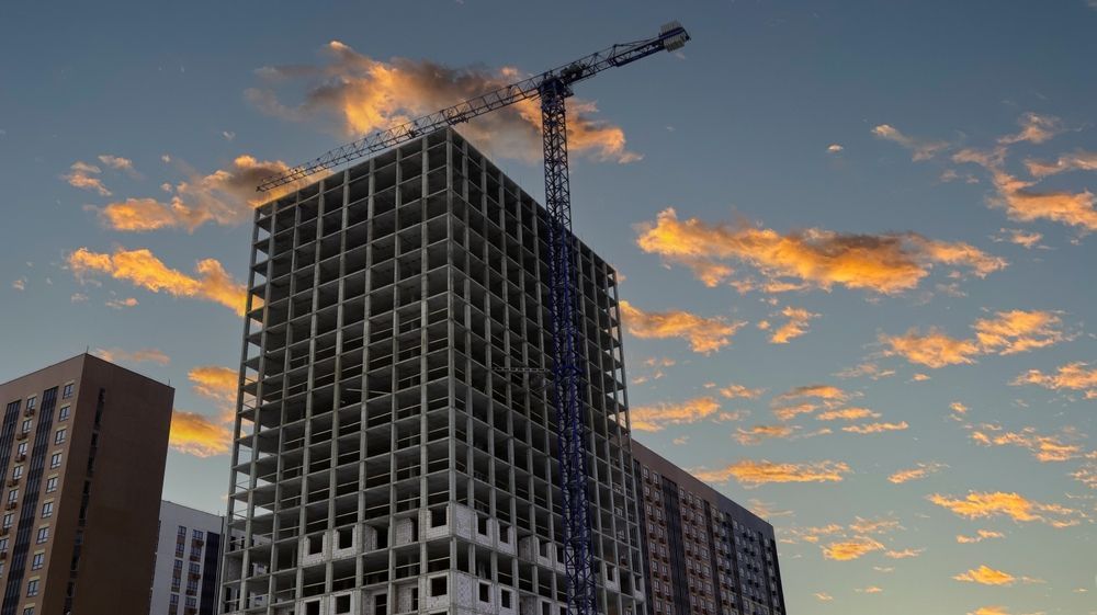 Construction of a high-rise building with a crane against a sunset sky with orange and yellow clouds.