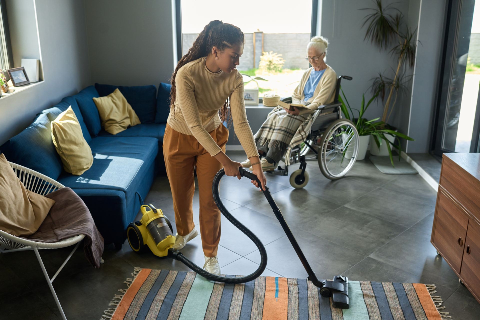 Woman vacuums a rug in a living room, while an elderly woman sits in a wheelchair nearby.